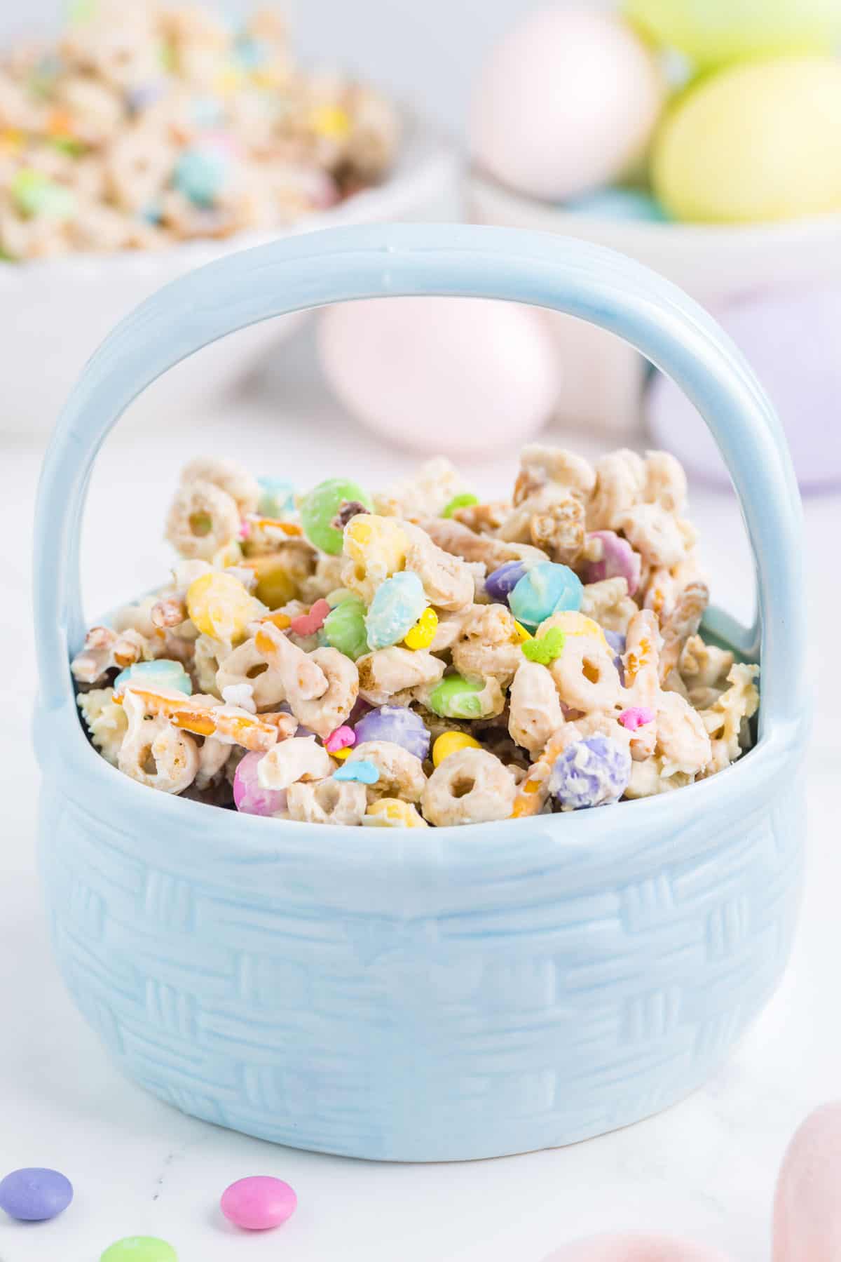 Easter bunny bait snack mix in a blue Easter basket dish with easter eggs and more bunny bait in the background and a few M&Ms around it