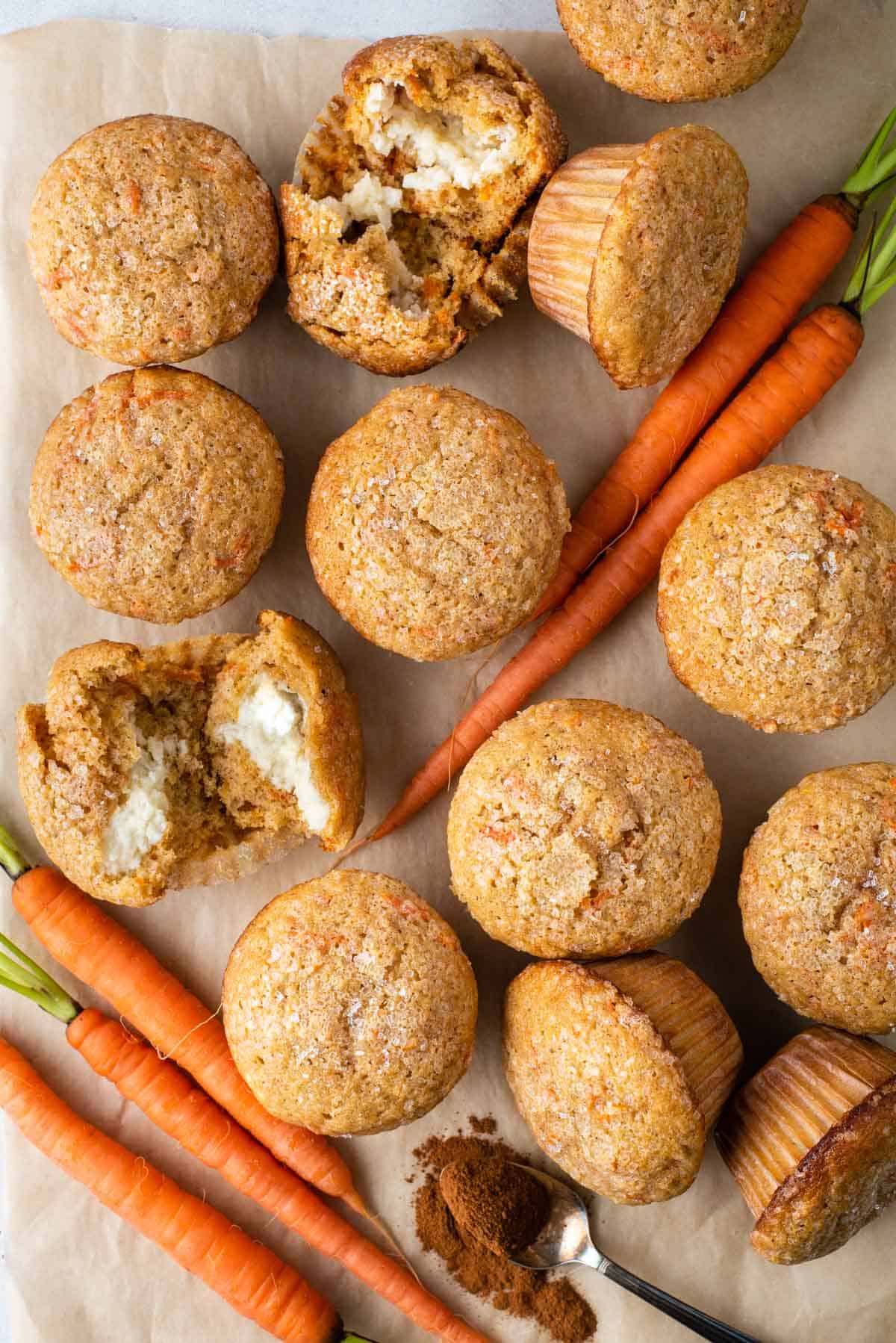carrot cake muffins arranged around carrots, a spoon of spices, and one muffin broken open to reveal the cream cheese filling inside