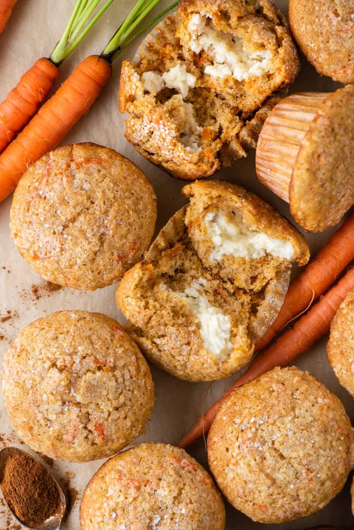 carrot cake muffins arranged around carrots, a spoon of spices, and one muffin broken open to reveal the cream cheese filling inside
