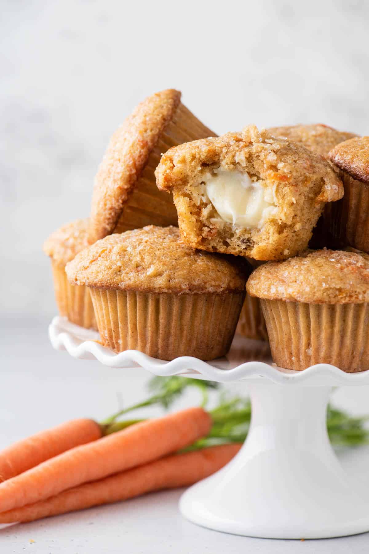 a pile of carrot cake muffins sitting on a white cake platter with carrots beneath it and one of the muffins half eaten, revealing the cream cheese filing inside