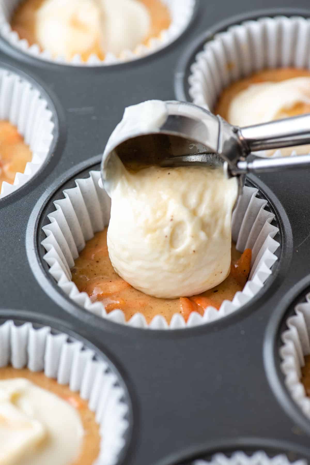 carrot cake muffin batter in the bottom of a paper liner in a muffin pan with cream cheese filling being placed on top of it