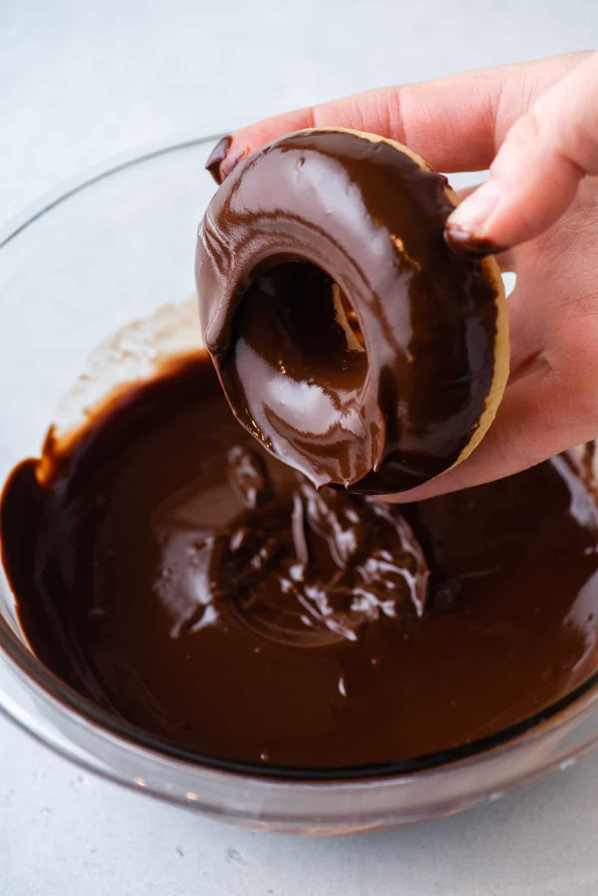 chocolate glaze for donuts in a clear glass bowl with a donut that has just been dipped in the glass being held above the bowl
