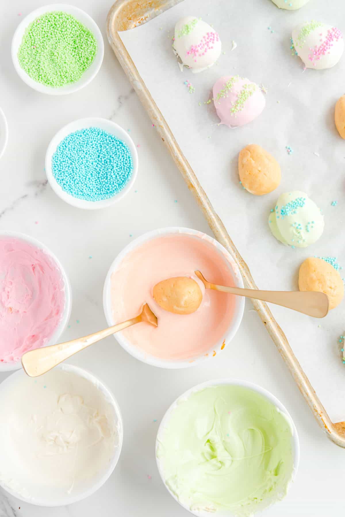a baking sheet with some dipped and some non-dipped easter egg truffles beside bowls of blue and green sprinkles, and bowls of pink, light orange, light green and white chocolate, with one truffle being dipped with forks into the light orange chocolate.