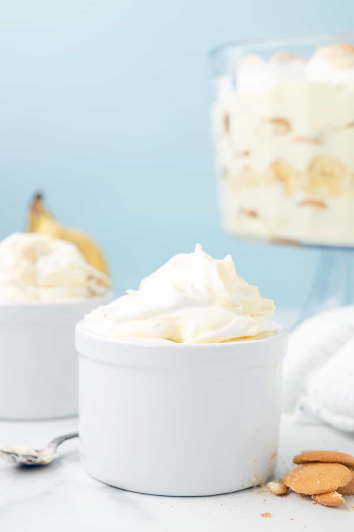 A white dish full of banana pudding arranged with vanilla wafers and a spoon around it with another dish and a trifle full of banana pudding in the background