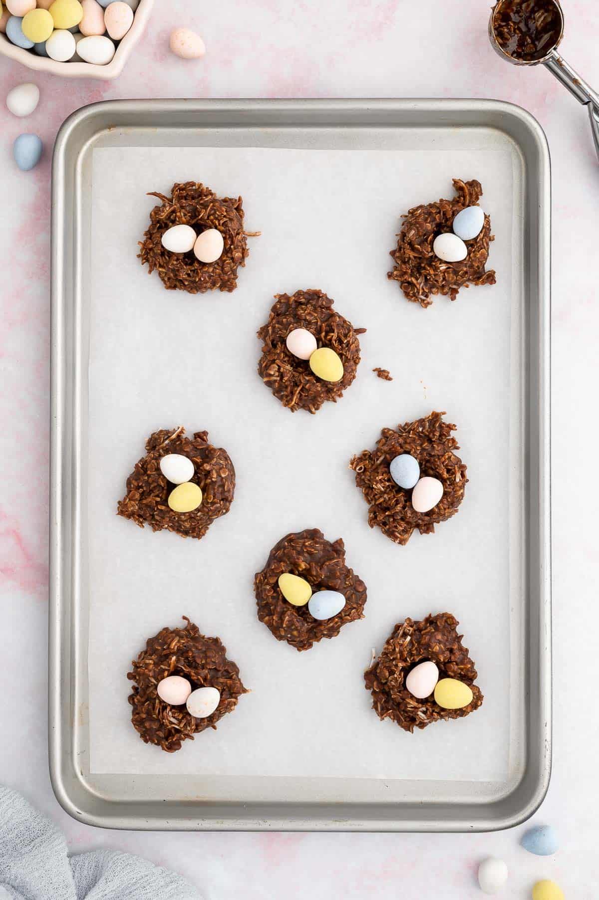 no bake birds nest cookies on a baking sheet surrounded by candy easter eggs and a cookie scoop