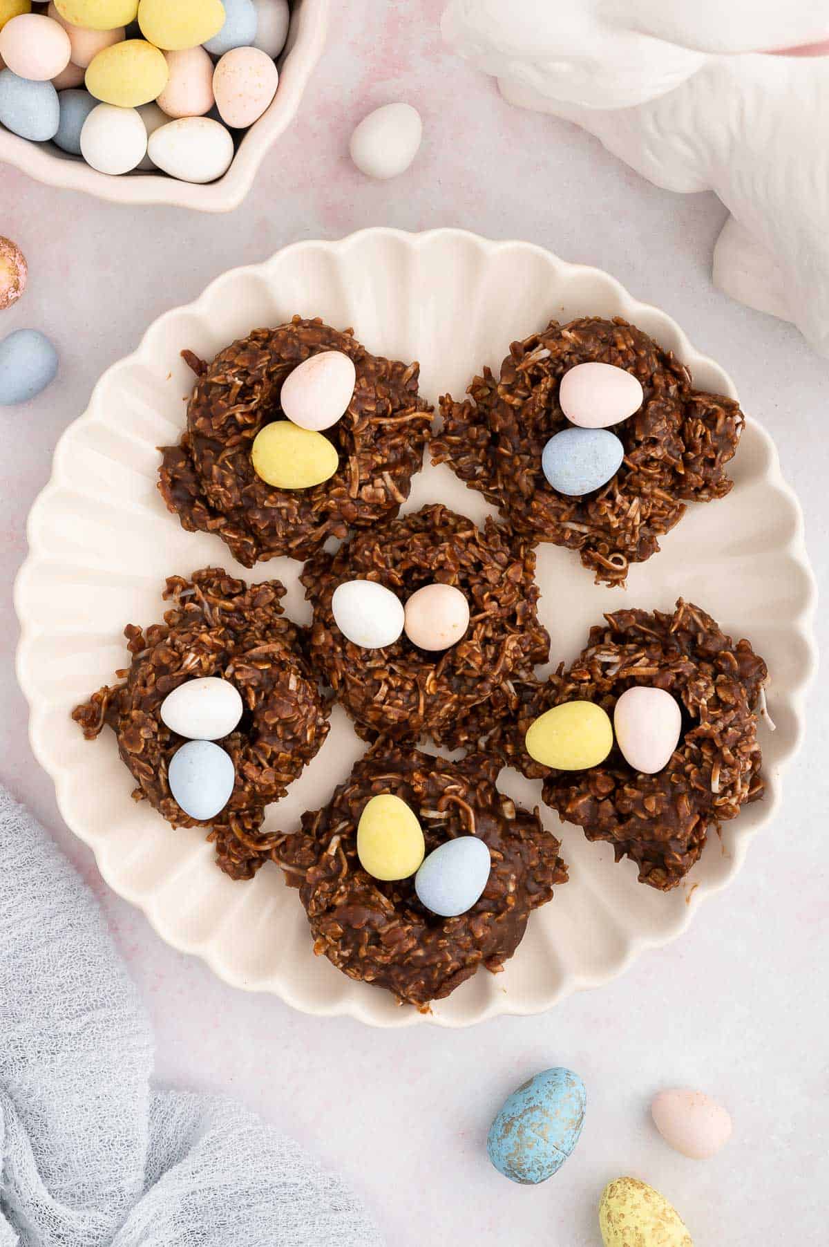 a plate of no bake birds nest cookies surrounded by a bowl of easter egg candies, a white ceramic bunny and more easter eggs