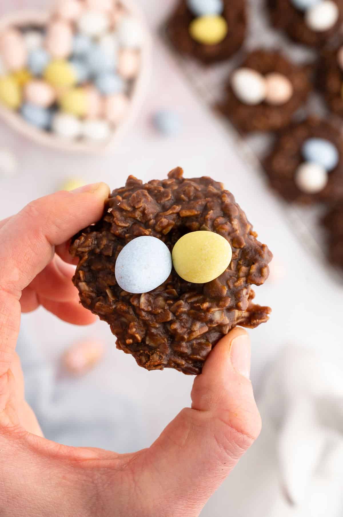 birds nest cookie being held up above a background of more cookies and a bowl of candy easter eggs