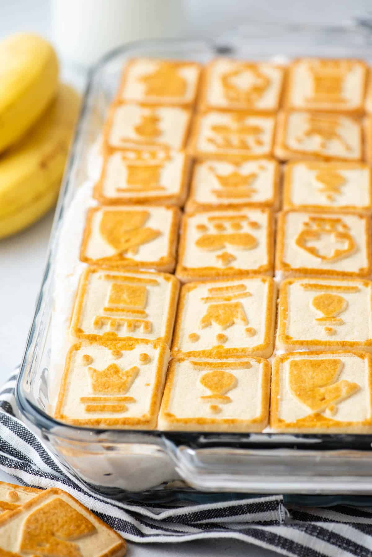 close up of Paula Deen banana pudding in a clear dish sitting on top of a black and white striped kitchen towel with chessman cookies around it