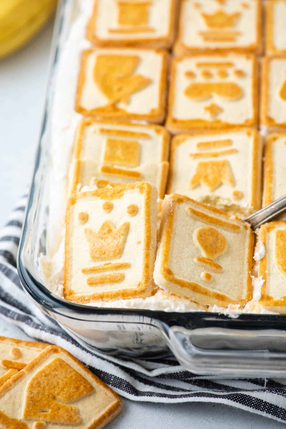 close up of Paula Deen banana pudding in a clear dish sitting on top of a black and white striped kitchen towel with chessman cookies around it