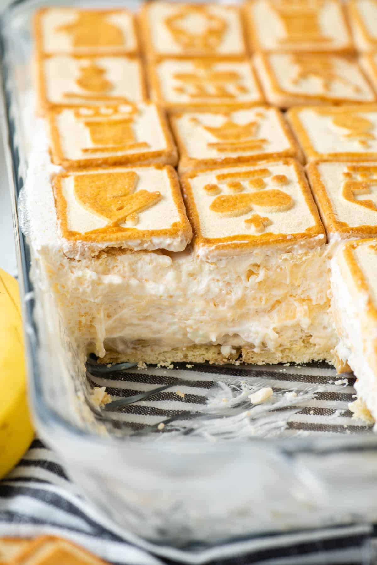 close up of partially eaten Paula Deen banana pudding in a clear dish sitting on top of a black and white striped kitchen towel with chessman cookies around it