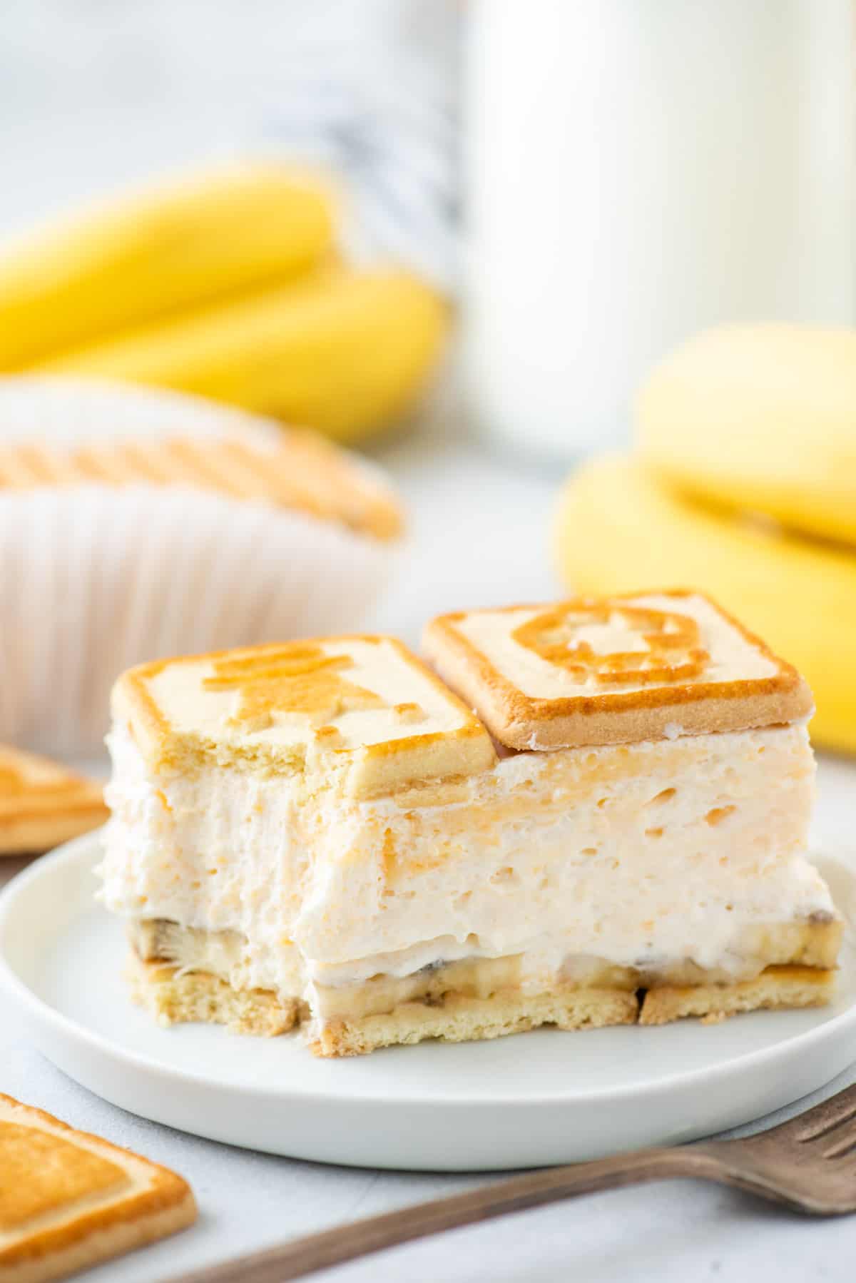 a serving of Paula Deen banana pudding on a white plate with a fork beside it arranged with bananas and more cookies around it