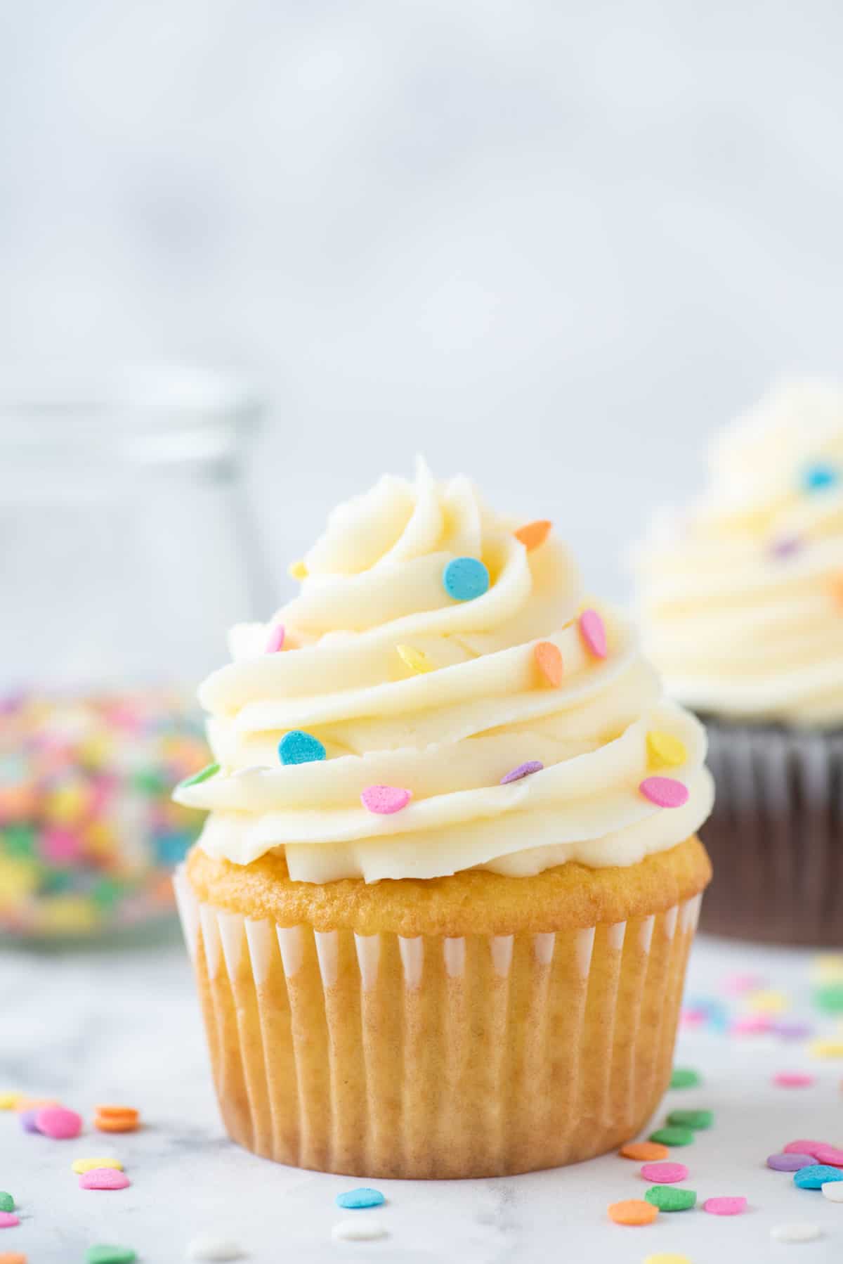 vanilla cupcake topped with vanilla frosting and sprinkles, with sprinkles all around it and more sprinkles and a chocolate cupcake in the background