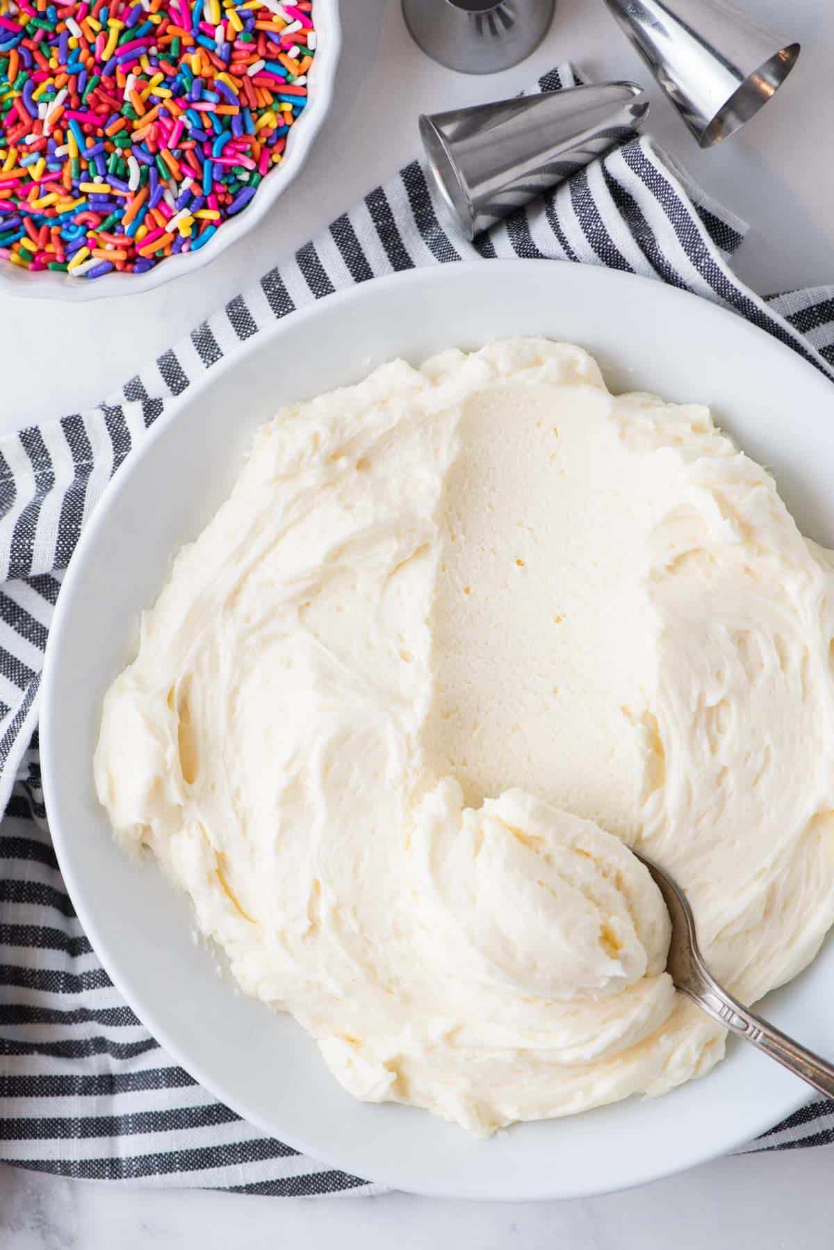 vanilla buttercream frosting in a white bowl sitting on top of a striped kitchen towel with a bowl of sprinkles and metal piping tips around it