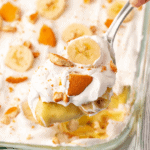 A close-up of an easy banana pudding recipe being scooped from a glass dish. The creamy dessert is topped with whipped cream, fresh banana slices, and crumbled vanilla wafers, making it irresistibly delightful.