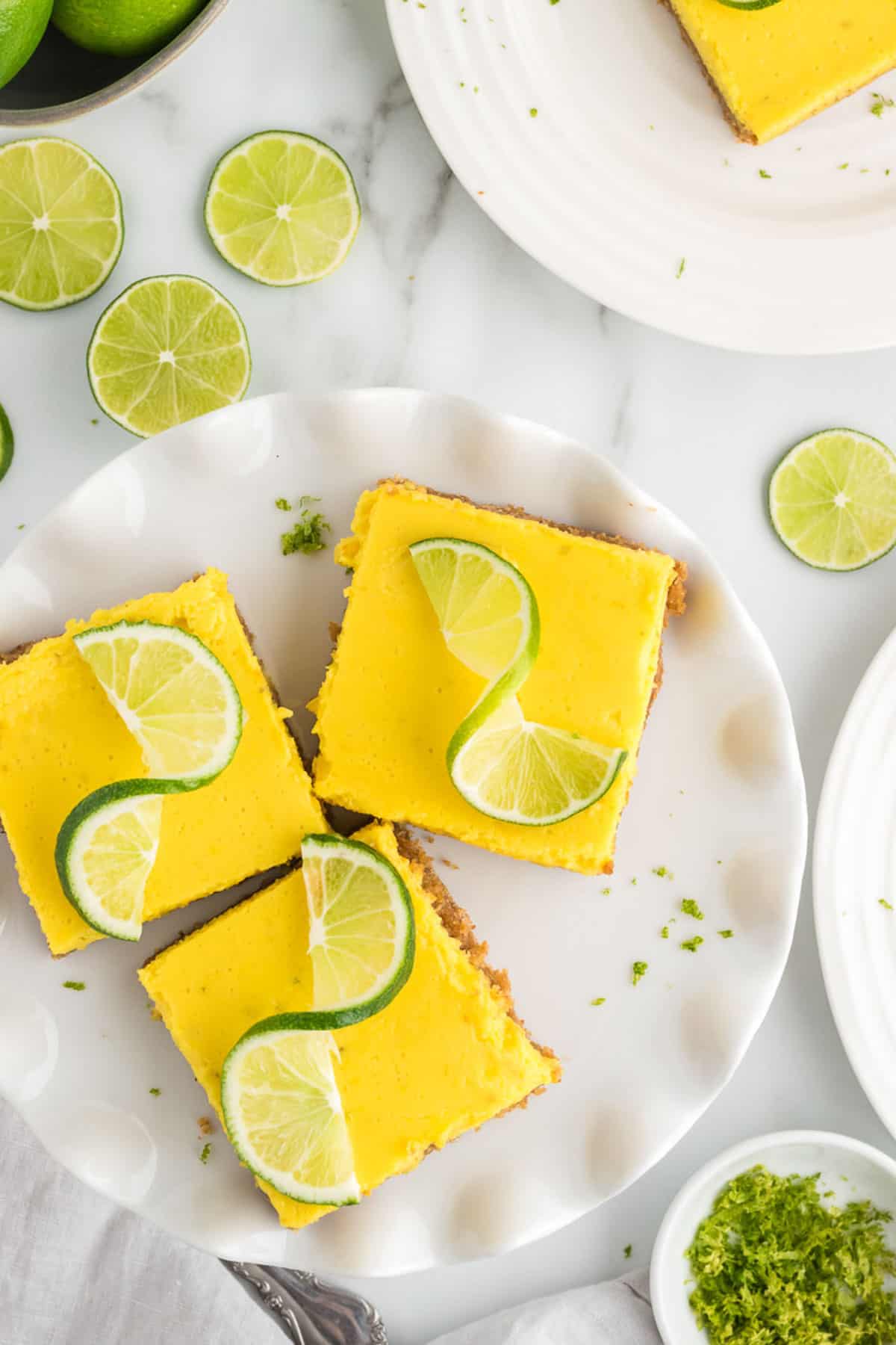 3 square key lime bars on a plate topped with fresh lime slices and sprinkled with lime zest, surrounded by a bowl of limes, another plate with a key lime bar on it more lime slices and a bowl of lime zest