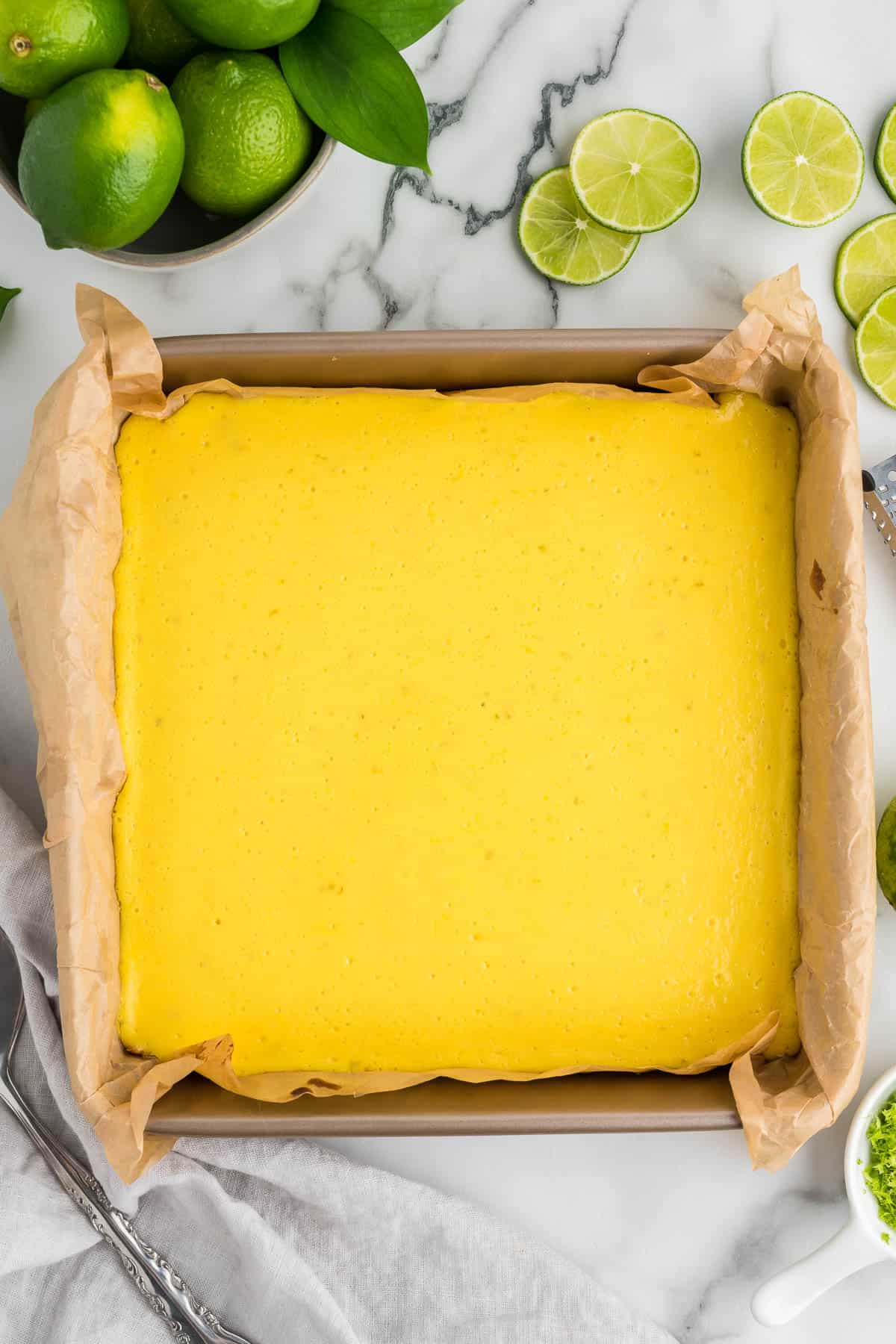 key lime bars in a square baking pan lined with parchment paper surrounded by a bowl of whole limes and fresh lime slices on the counter beside the pan