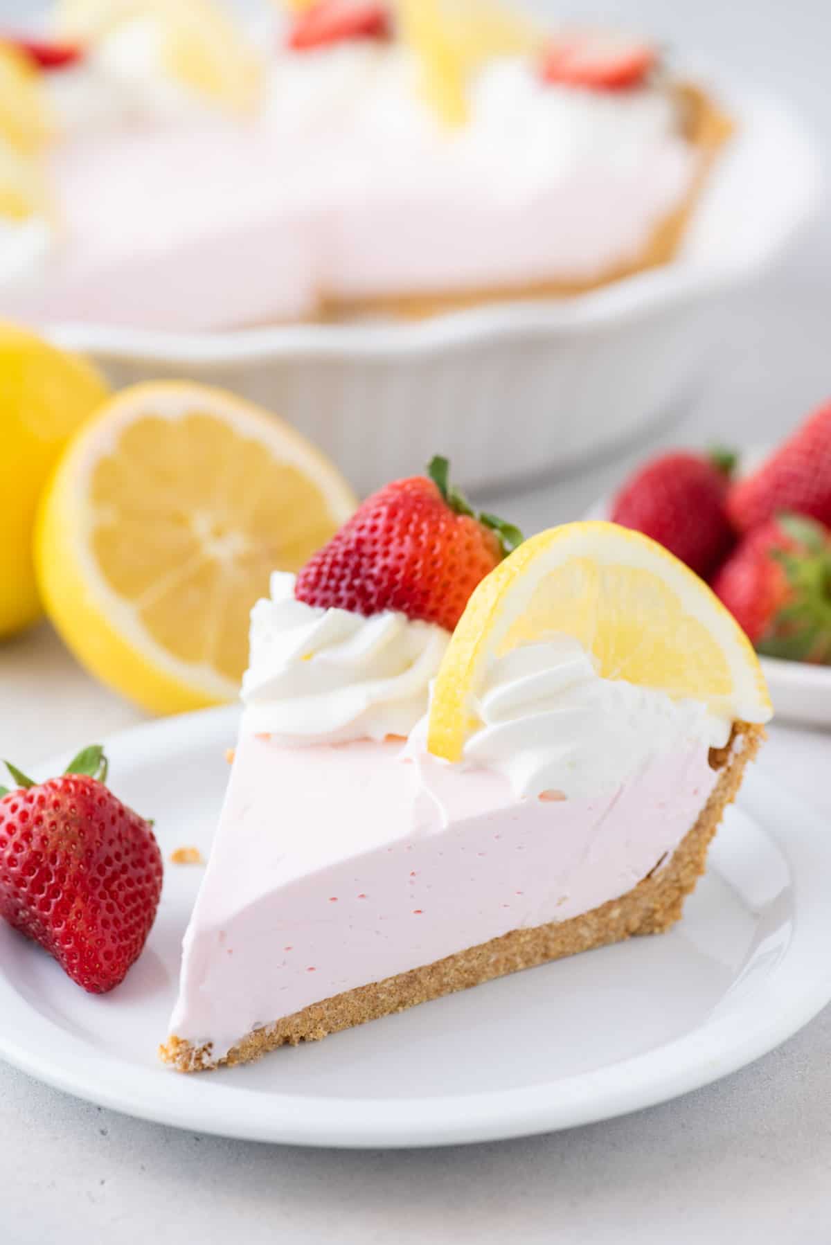 a slice of kool aid pie on a white plate surrounded by fresh strawberries, lemons and more pie in the background