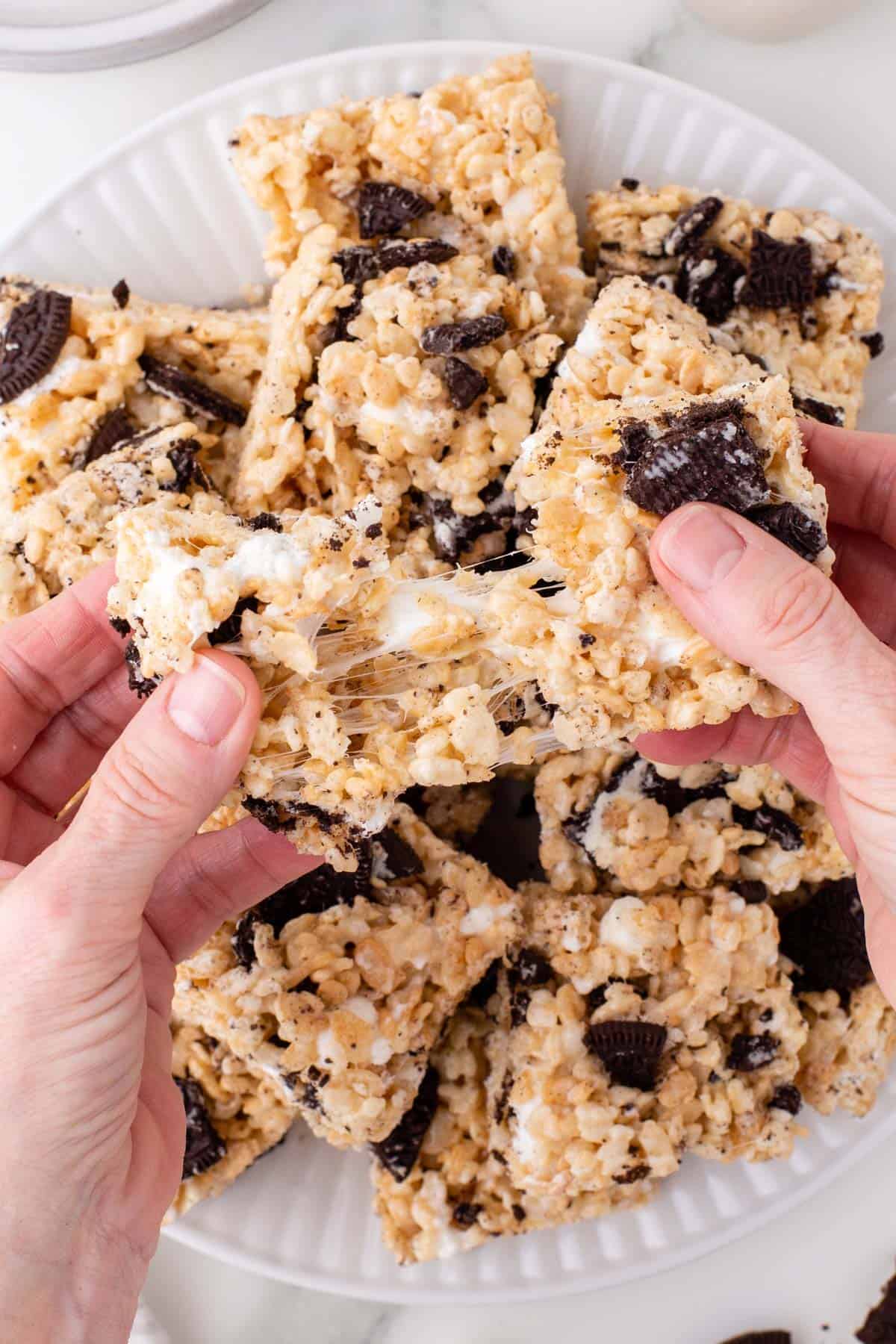 two hands pulling apart a gooey oreo rice krispies treat above a plate full of more rice krispie treats