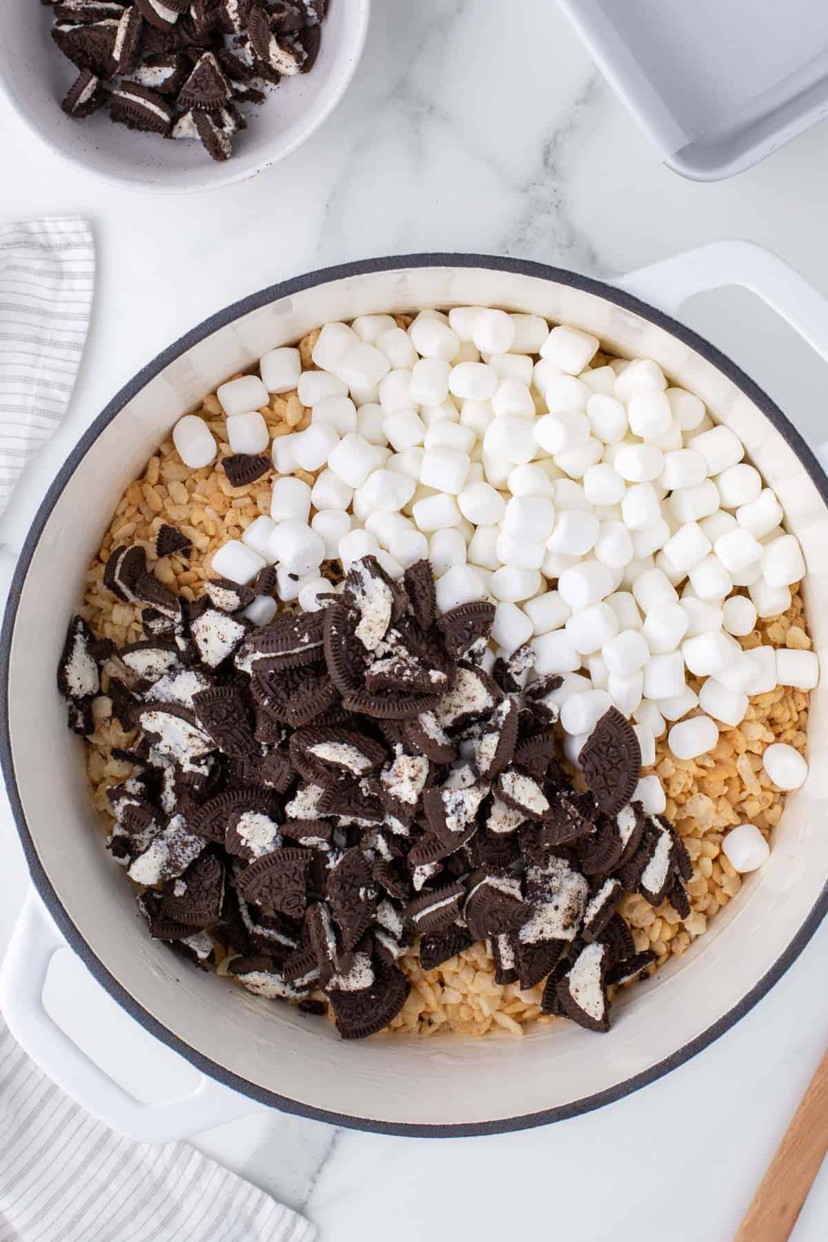 crumbled oreo cookies, mini marshmallows and rice krispie cereal in a white pot surrounded by a kitchen town, wooden spatula and a bowl of more crumbled oreos.