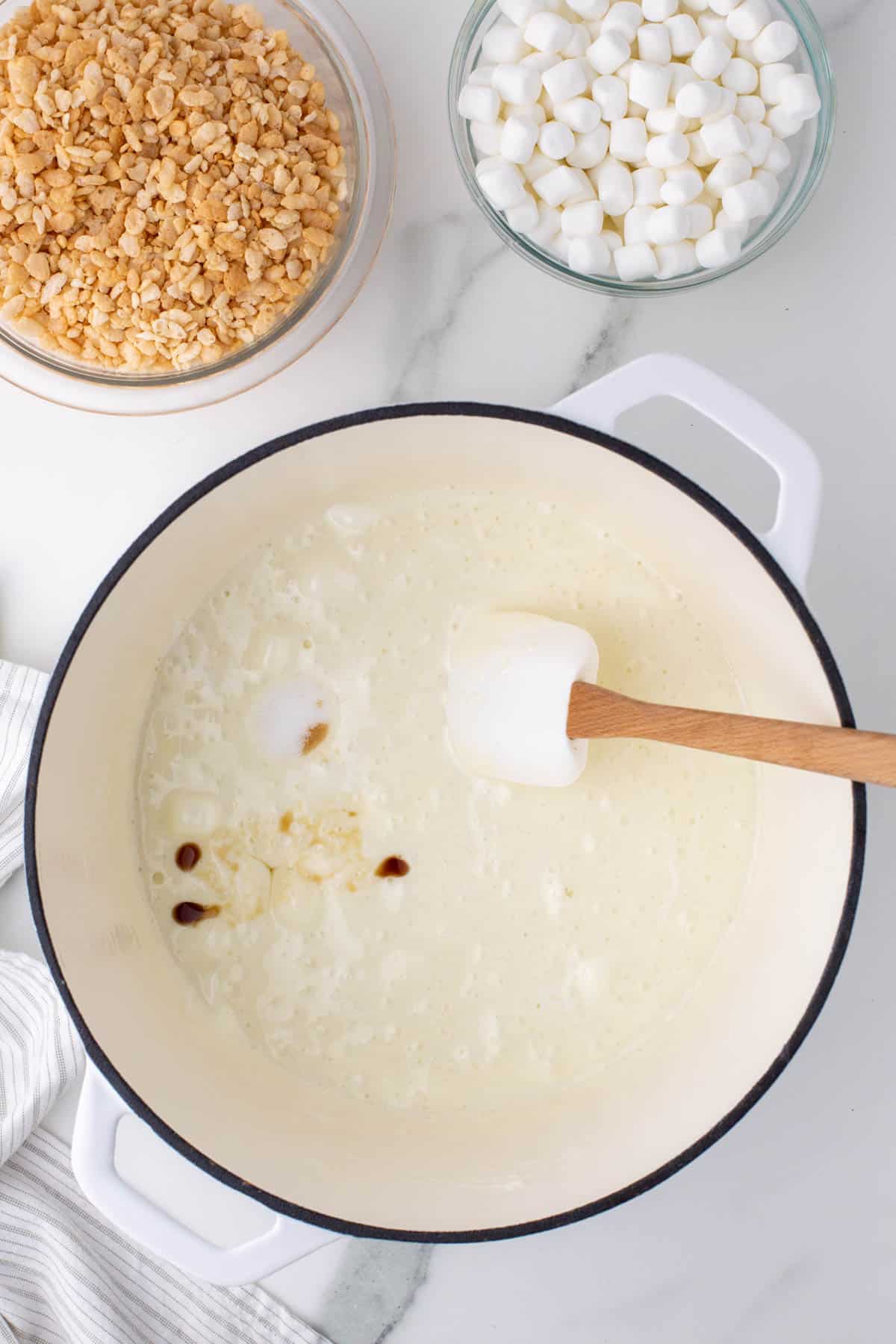 melted mini marshmallows in a white pot surrounded by a bowl of rice krispies, a bowl of mini marshmallows and a kitchen towel