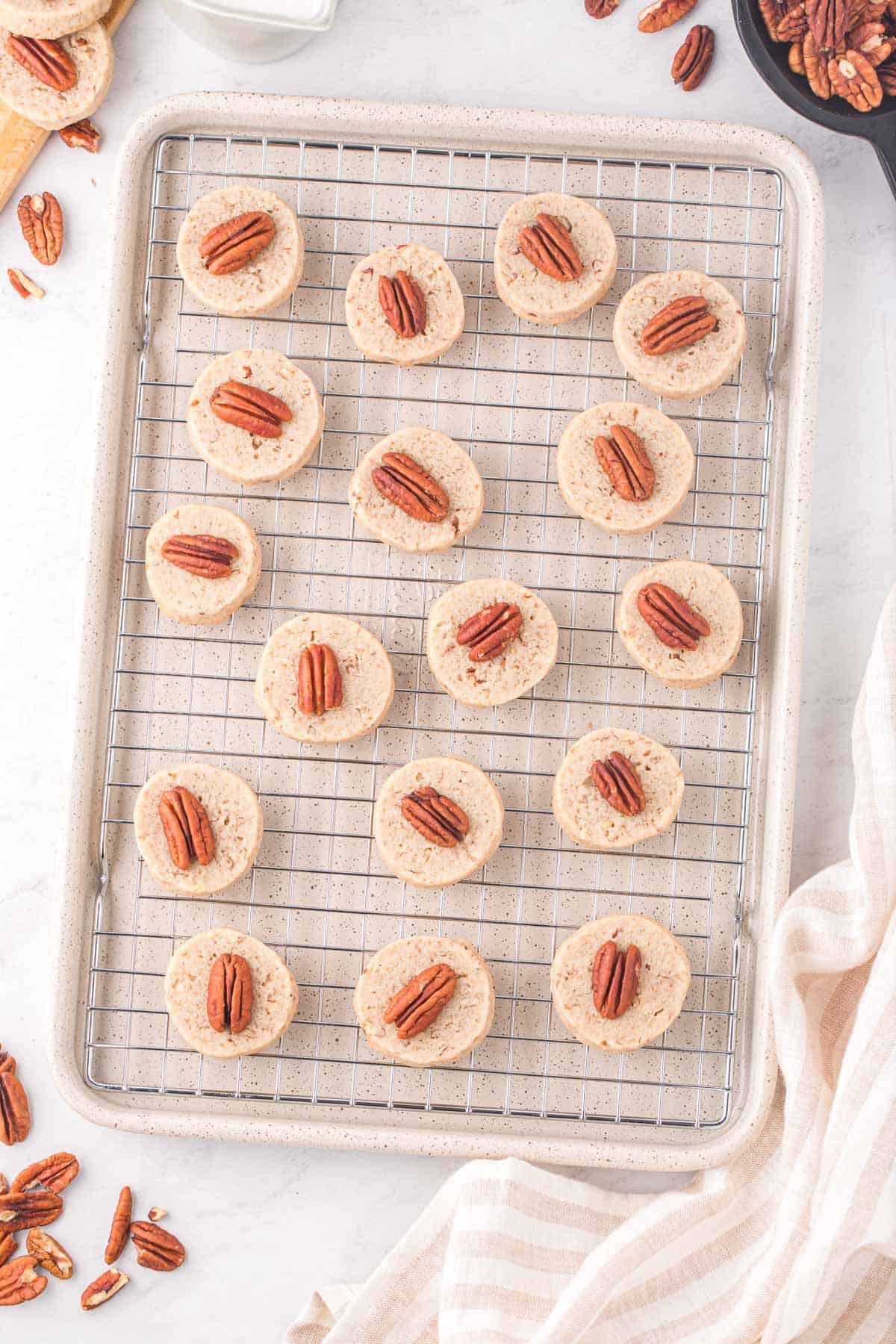 pecan sandies  lined in rows on a wire rack on top of a backing dish surrounded by a kitchen towel, more pecan pieces and more cookies