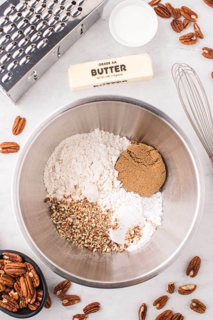 dry ingredients for pecan sandies in a mixing bowl surrounded by a stick of butter, pecans, a grater, and a whisk