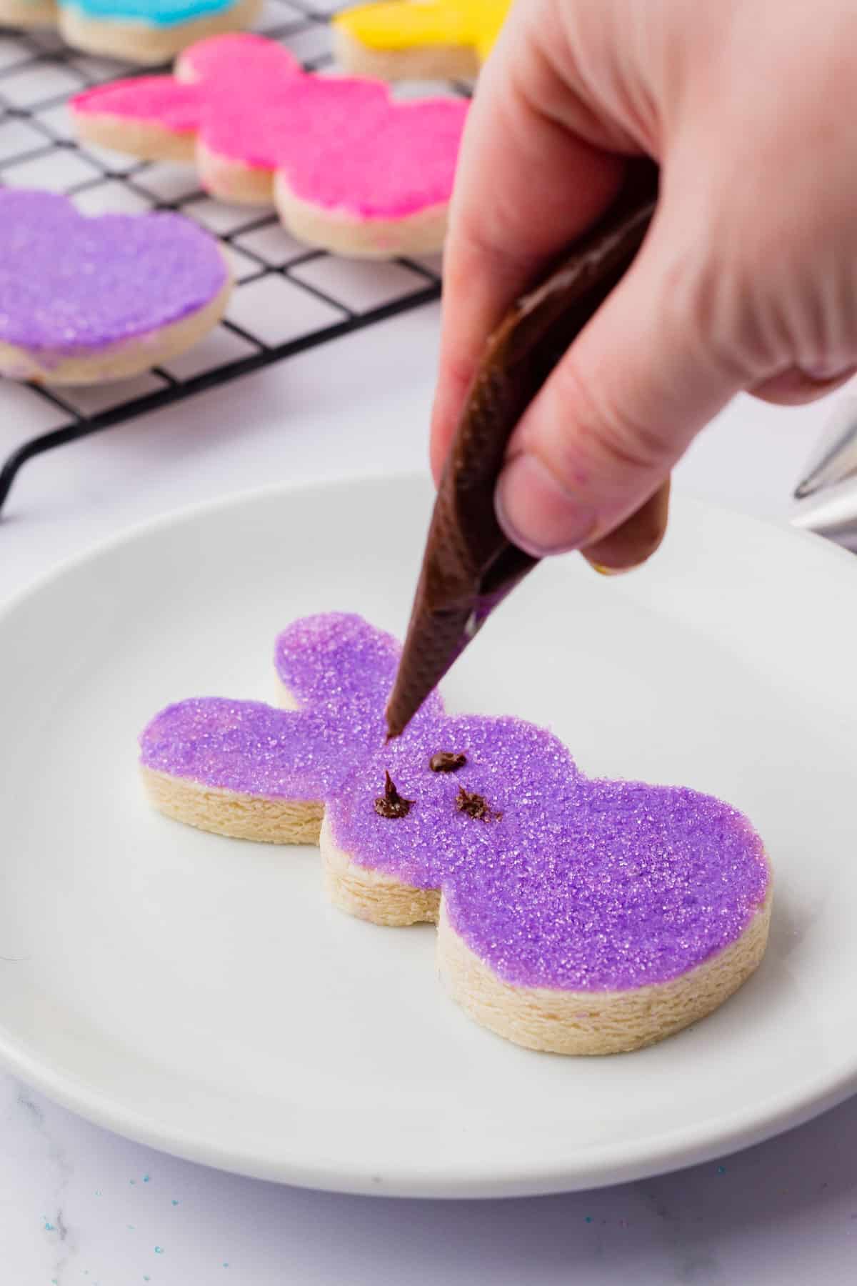 Peeps Bunny sugar cookie on a white plate with purple frosting and sugar on top being decorated with chocolate eyes and a nose
