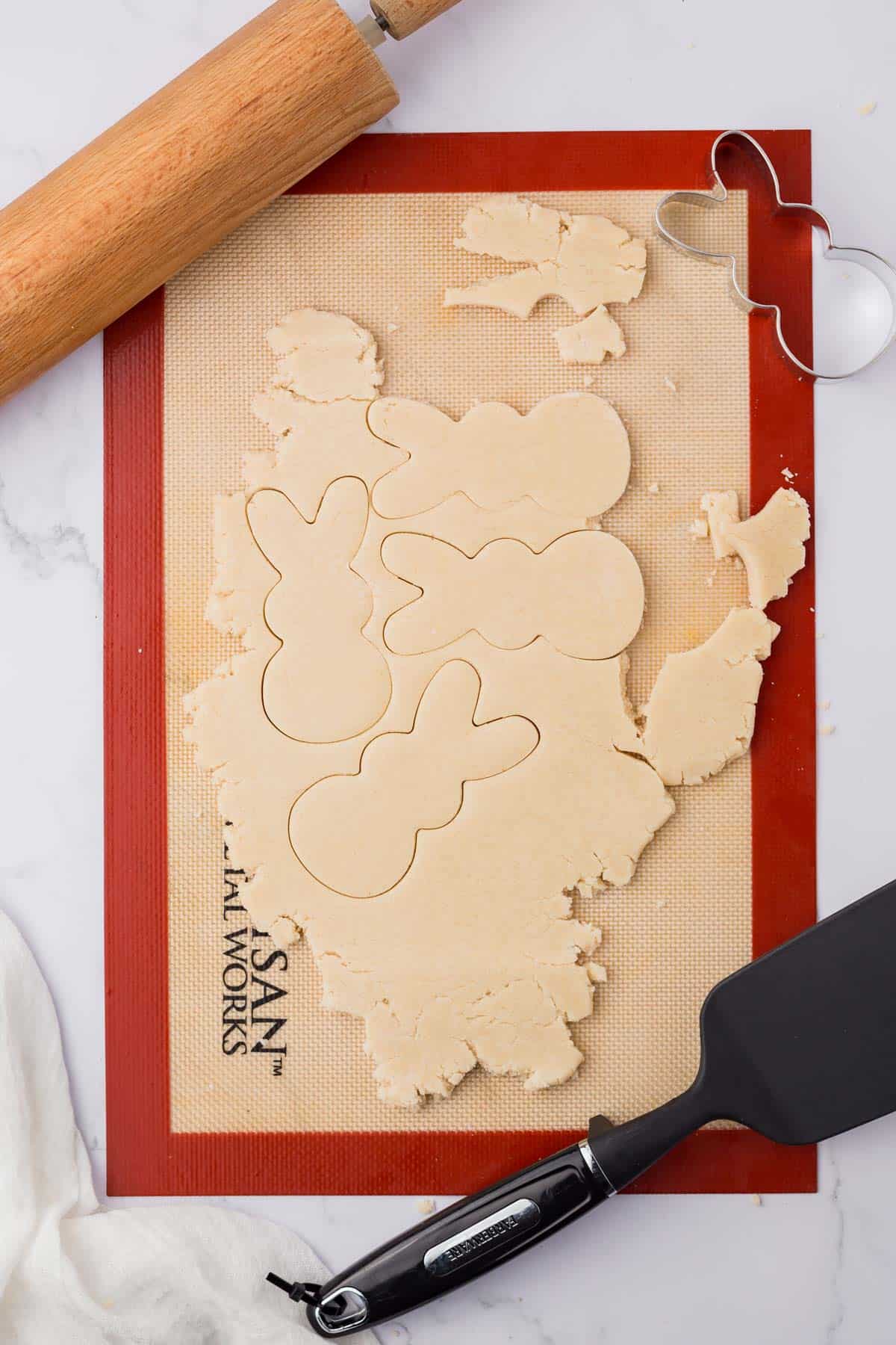 silicone mat with sugar cookie dough on top, with bunny cutout prints in it, surrounded by a rolling pin, spatula, and peeps bunny cookie cutter