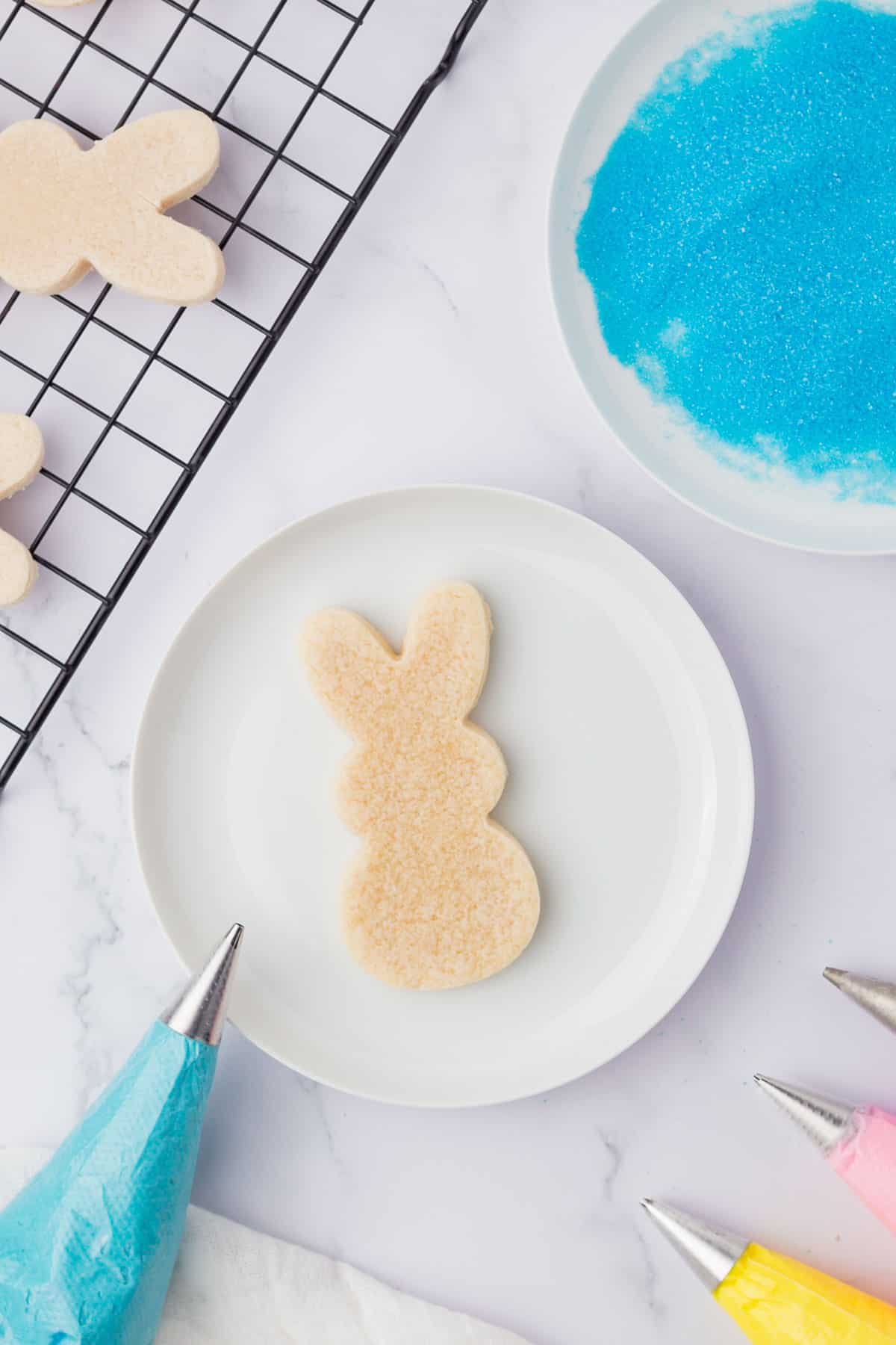 an undecorated bunny sugar cookie on a white plate surrounded by a bowl of blue sugar, piping bags with colored icing and cooling rack with more cookies