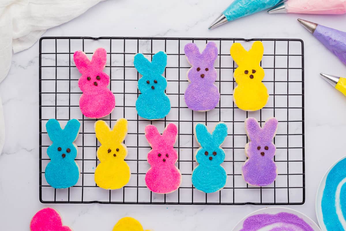 peeps bunny cookies lined up in two rows on a cooling rack, surrounded by piping bags with blue, pink, purple and yellow icing and bowls of purple and blue sugar