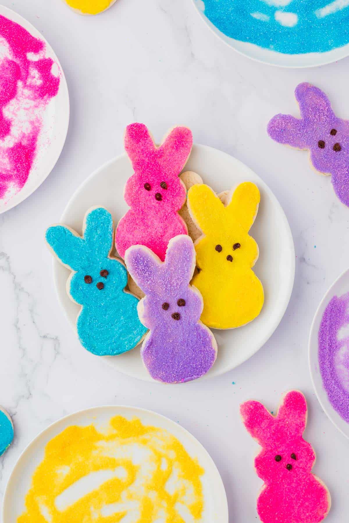 pink, yellow, blue and purple peeps bunny sugar cookies on a white plate, surrounded by bowls of colored sugar and more cookies