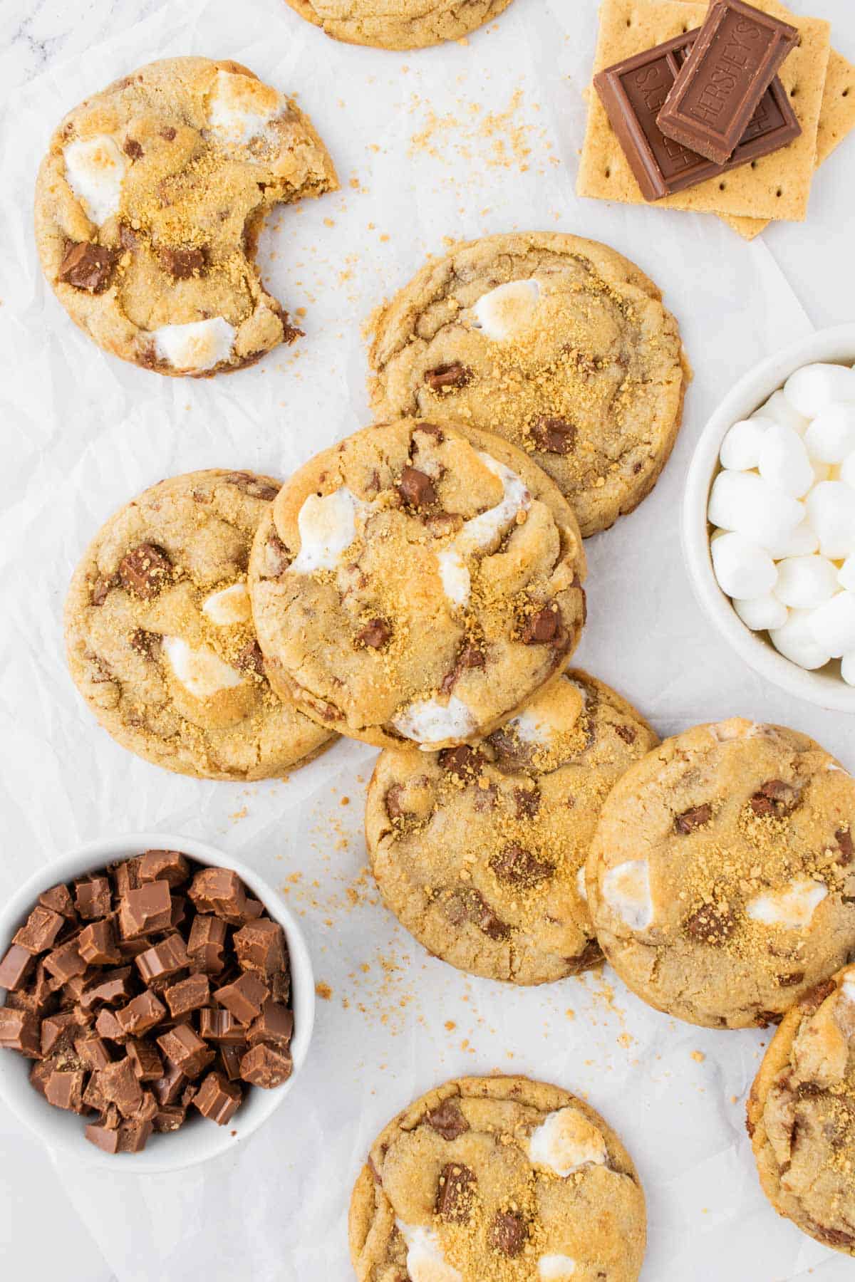 a pile of smores cookies with graham crackers, chocolate, marshmallows, and a partially eaten cookie arranged around them