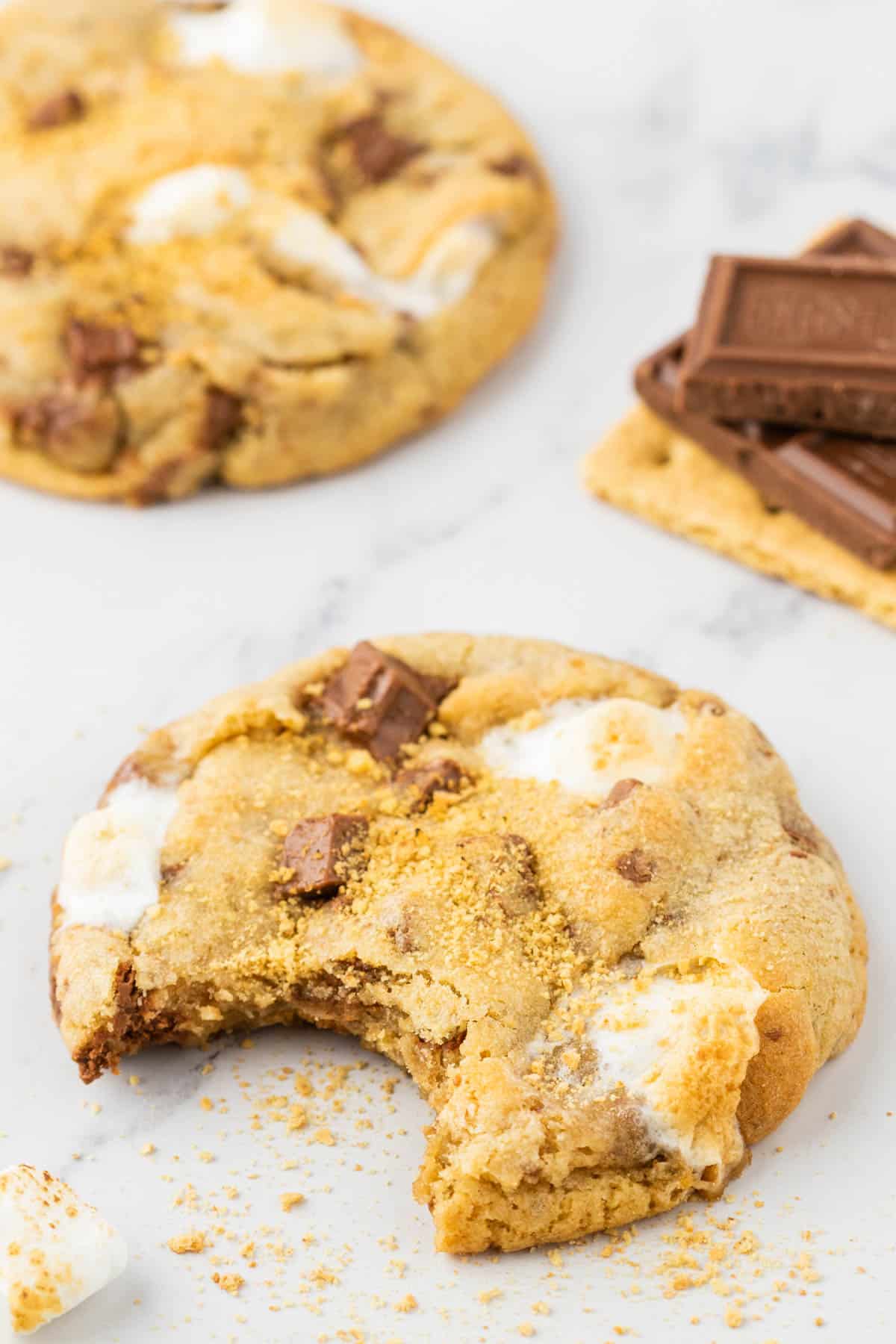 a smores cookie with a bite out of it with crumbs around it, another cookie in the background and a stack of graham cracker with hersheys chocolate on top