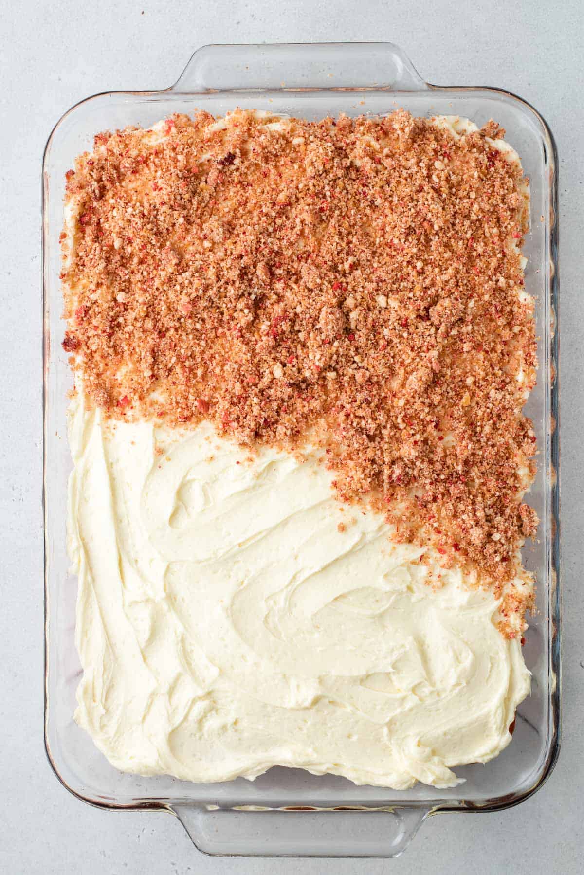 strawberry cake in a glass baking dish covered in vanilla frosting and half covered in strawberry crunch topping