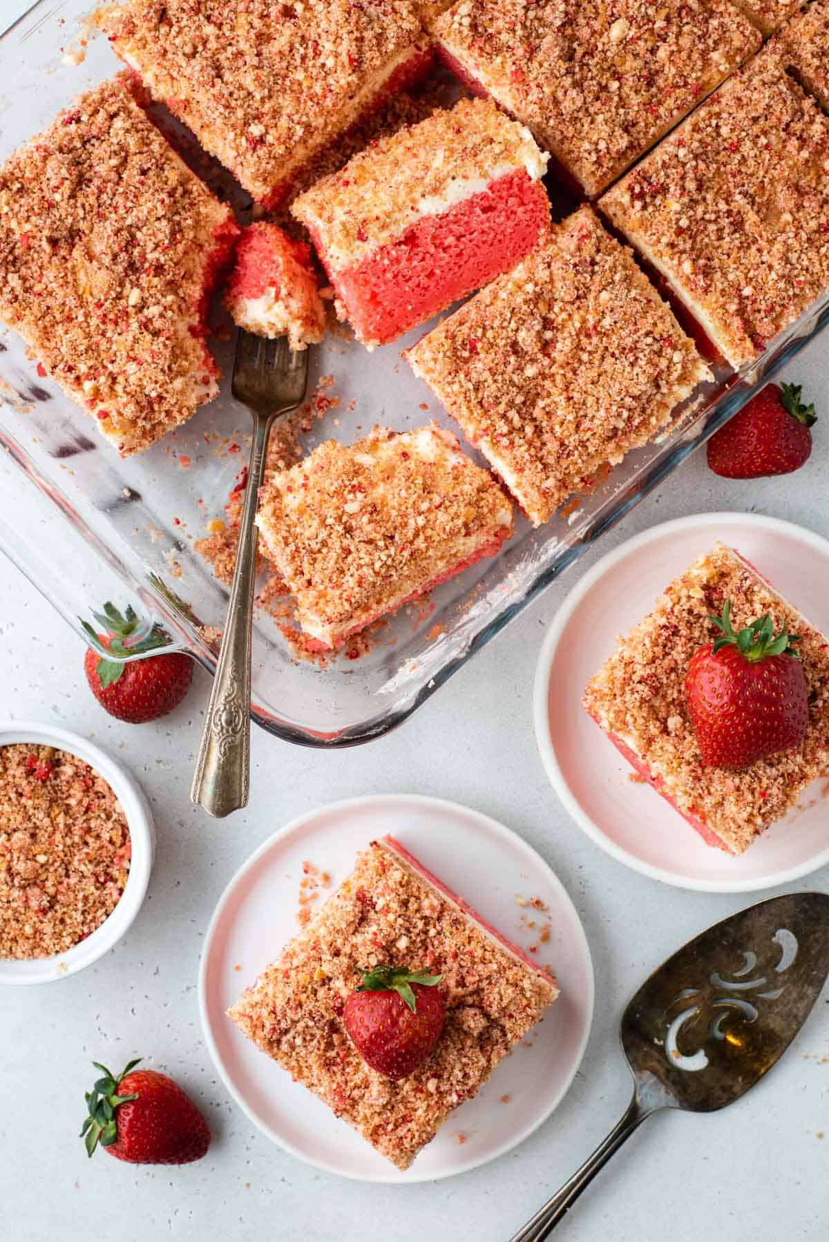 clear glass baking dish full of sliced strawberry crunch cake beside two white plates with slices of cake on them, strawberries, a spoon, and a bowl of cake crumble topping