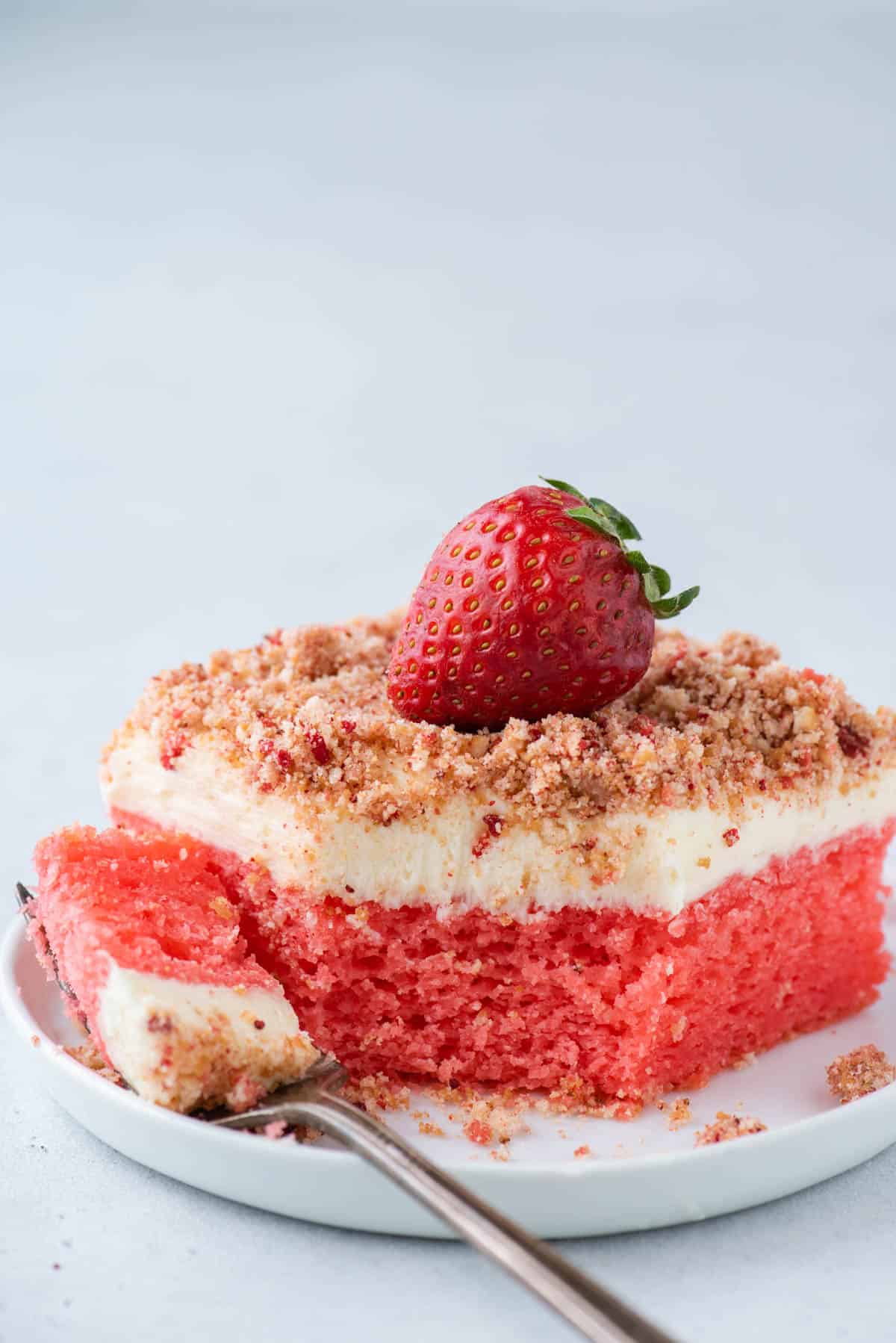 a slice of strawberry crunch cake on a white plate with a fork laying on the plate that has one bite of cake on it
