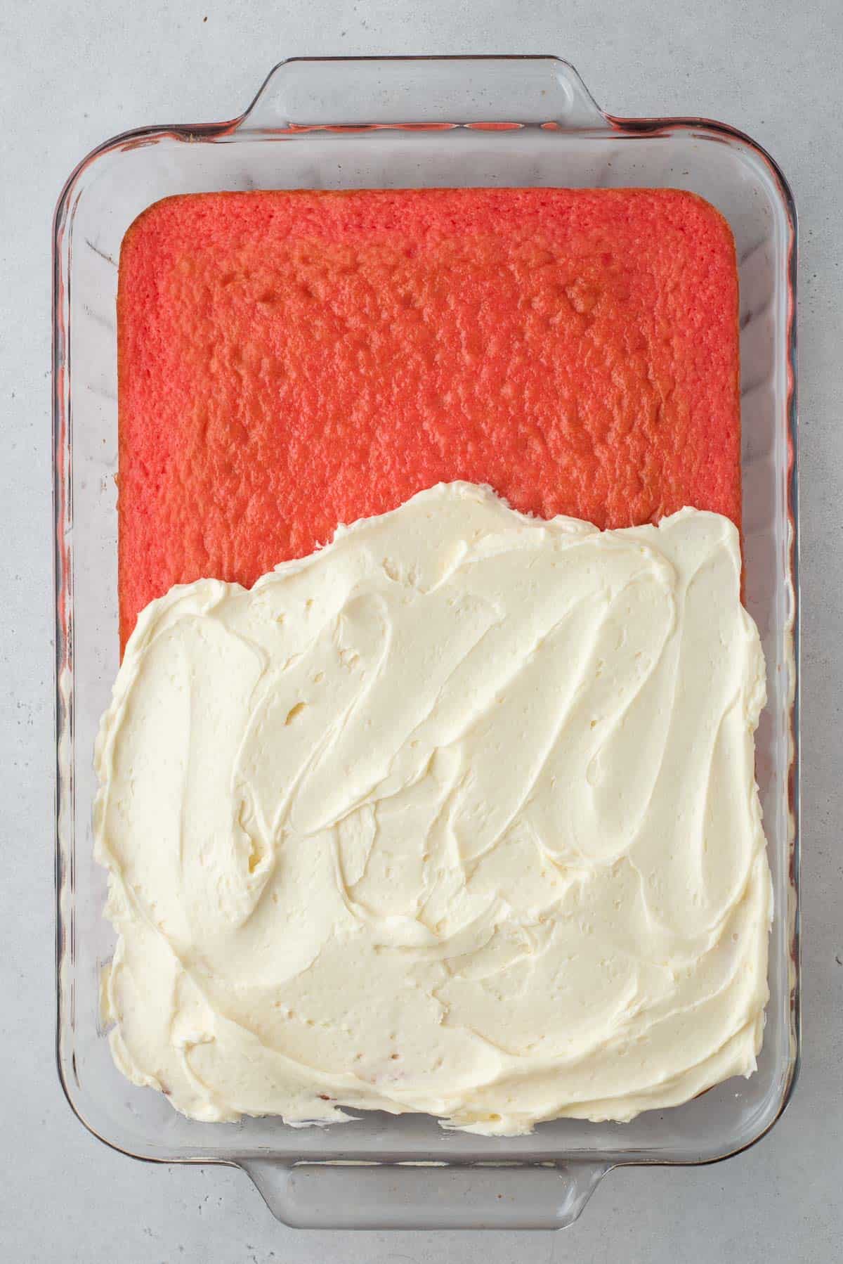 strawberry cake in a glass baking dish half covered in vanilla frosting