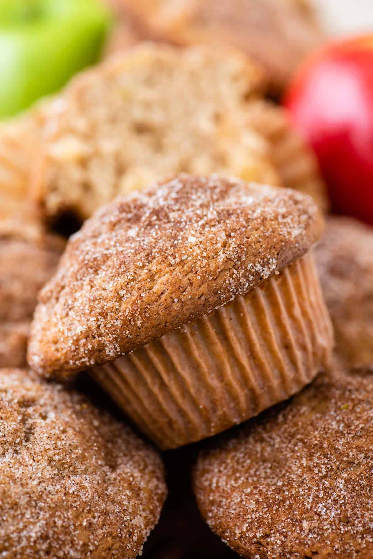 a pile of apple cinnamon muffins topped with cinnamon sugar with red and green apples in the background
