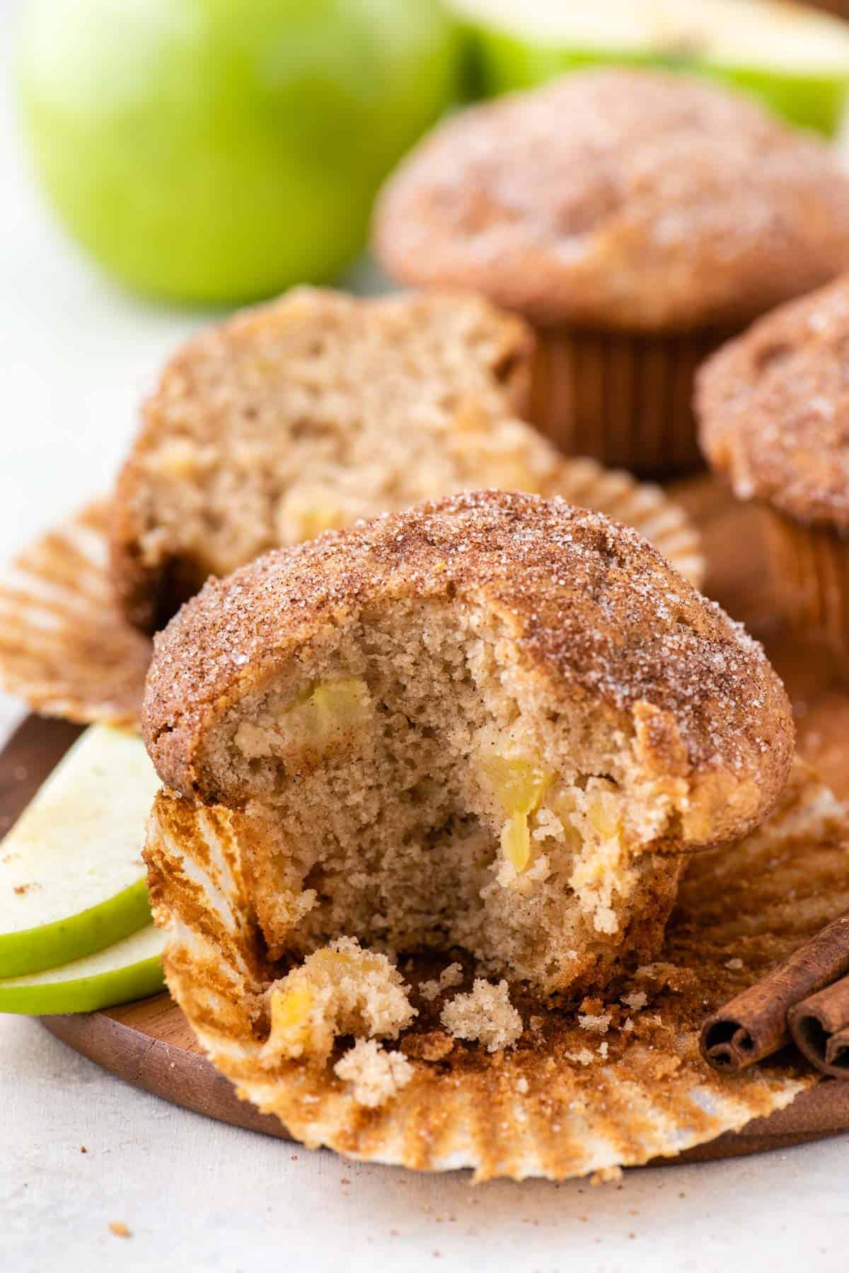 a partially eaten apple cinnamon muffin sitting inside its muffin paper surrounded by cinnamon sticks and green apple slices