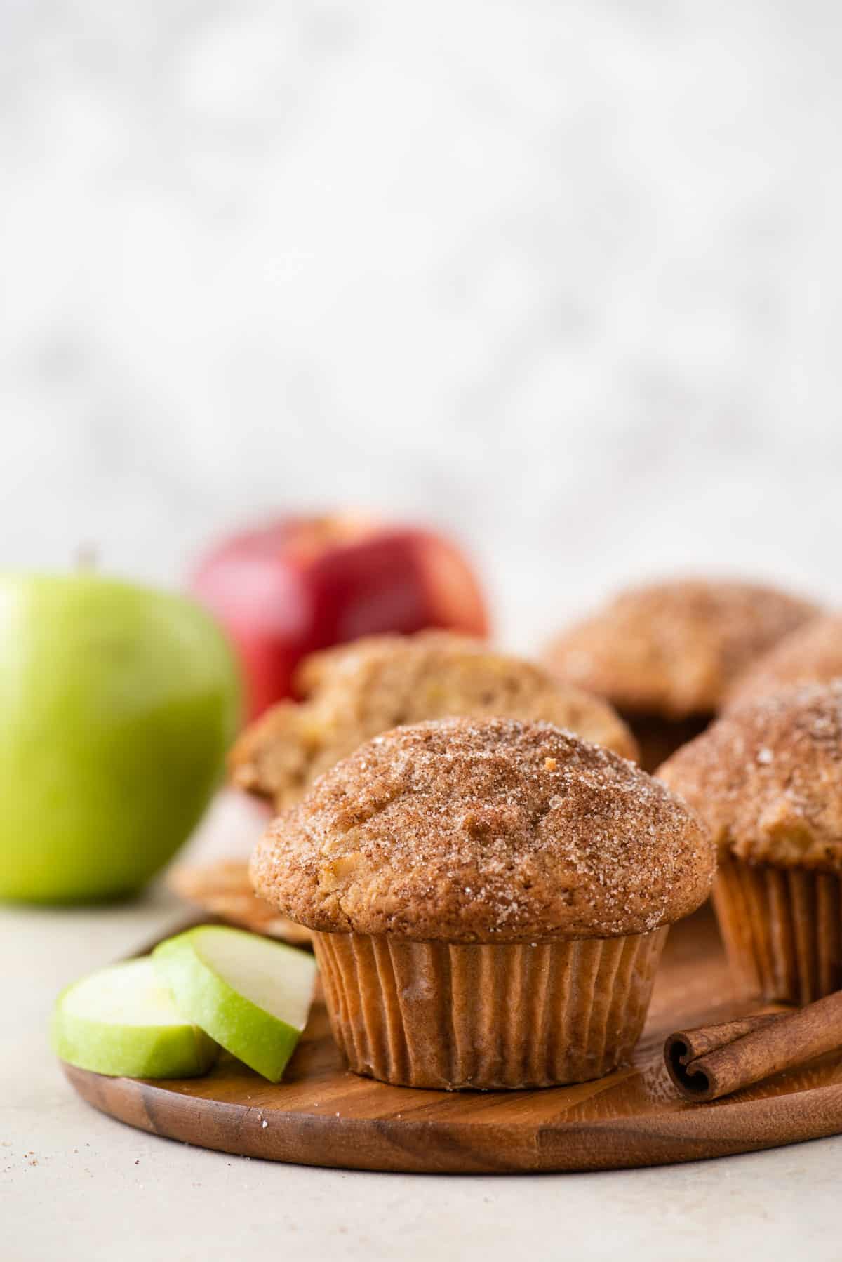 apple cinnamon muffins on a wood platter surrounded by a cinnamon stick, green apple slices, and more apples and muffins in the background
