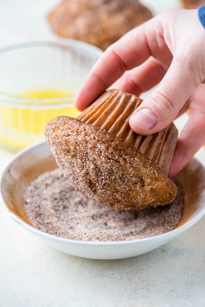 apple cinnamon muffin with the top being dipped into a bowl of cinnamon sugar with a dish of melted butter in the background