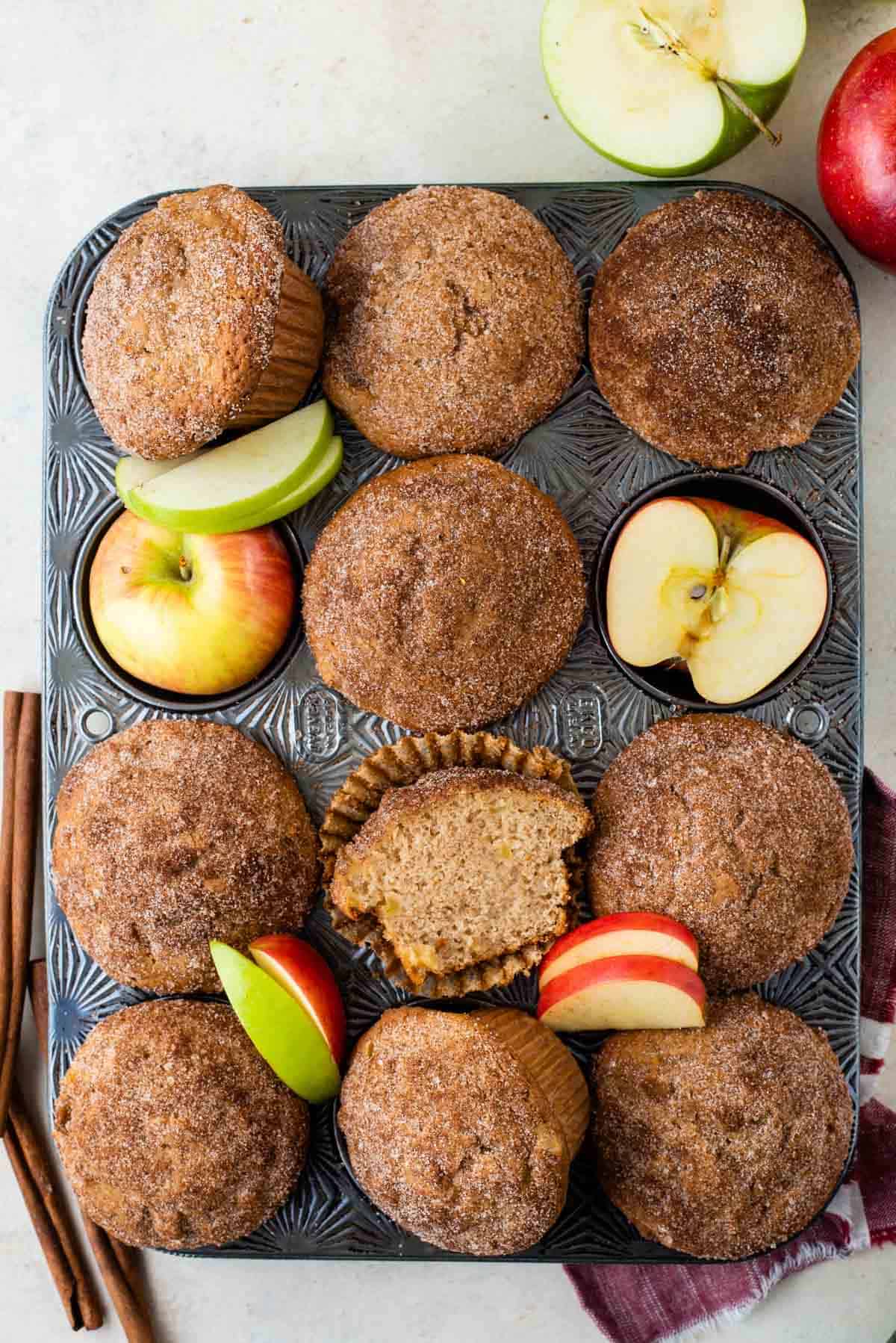 a muffin pan full of apple cinnamon muffins, red and green apple slices, half apples and whole apples, surrounded by cinnamon sticks and more apples