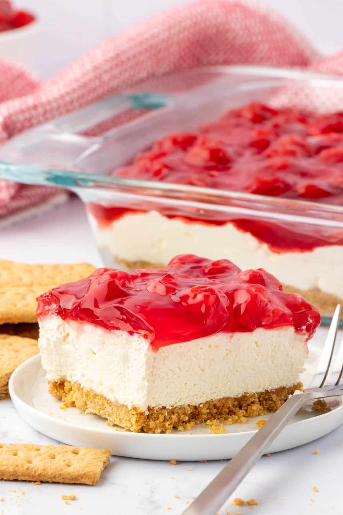 a slice of cherry delight on a white plate with a fork surrounded by graham crackers with a dish full of cherry delight and a red and white towel in the background