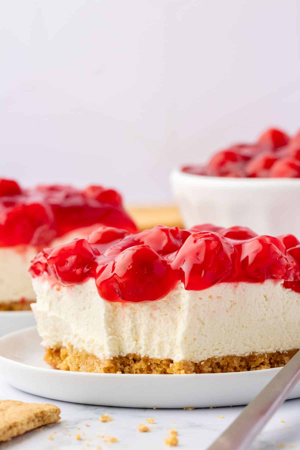 cherry delight on a white plate surrounded by graham crackers, another slice on a white plate, and a white bowl filled with cherry pie filling in the background