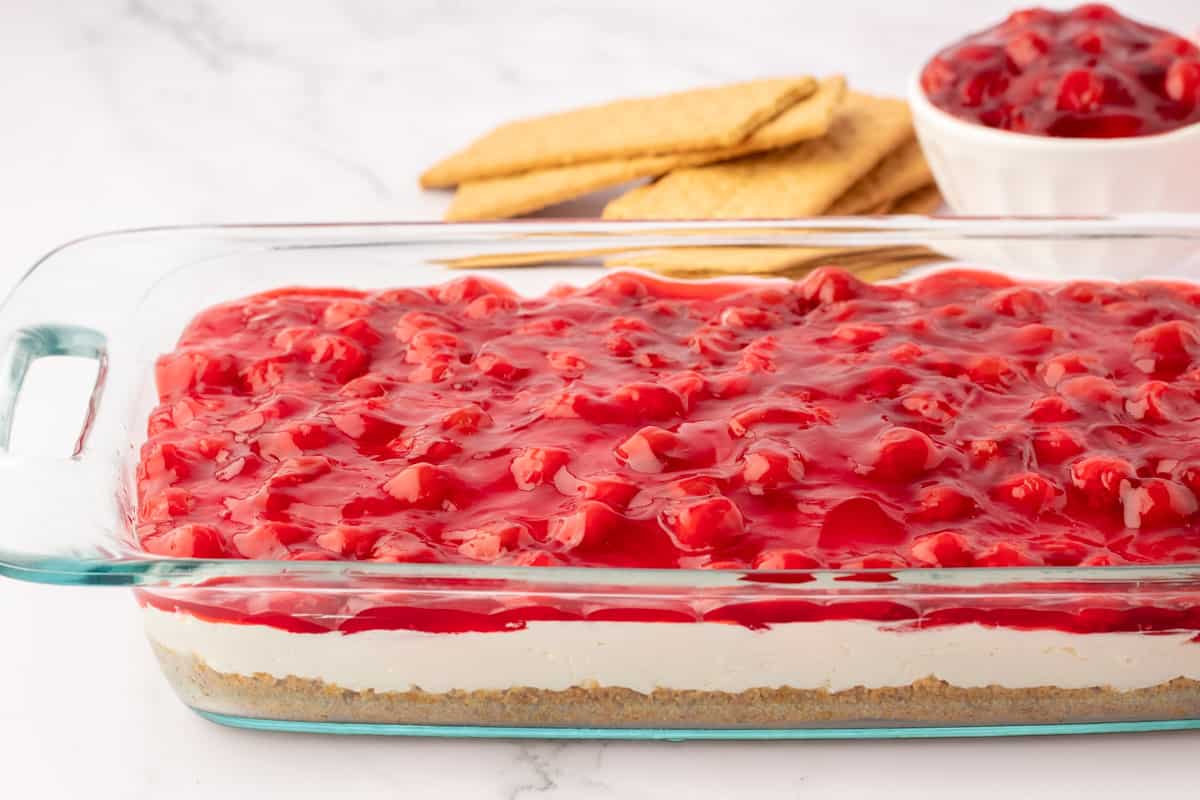 a clear baking dish showing 3 layers of graham cracker crust, cream cheese filling and cherry pie filling with graham crackers and a bowl of cherry pie filling in the background
