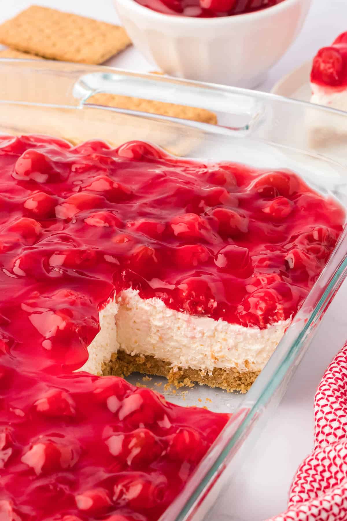 no bake cherry cheesecake in a clear baking dish with one slice cut out surrounded by a red and white towel, graham crackers, and a bowl of cherry pie filling