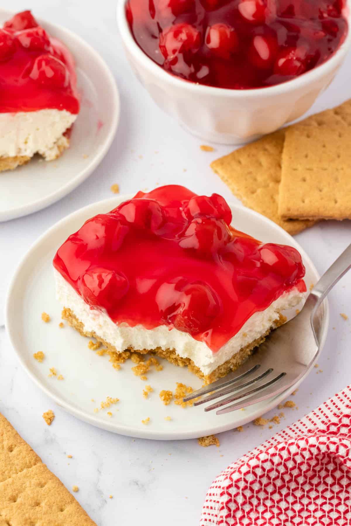 twice slices of cherry delight on white plates surrounded by graham crackers, a red and white towel, and a white bowl filled with cherry pie filling