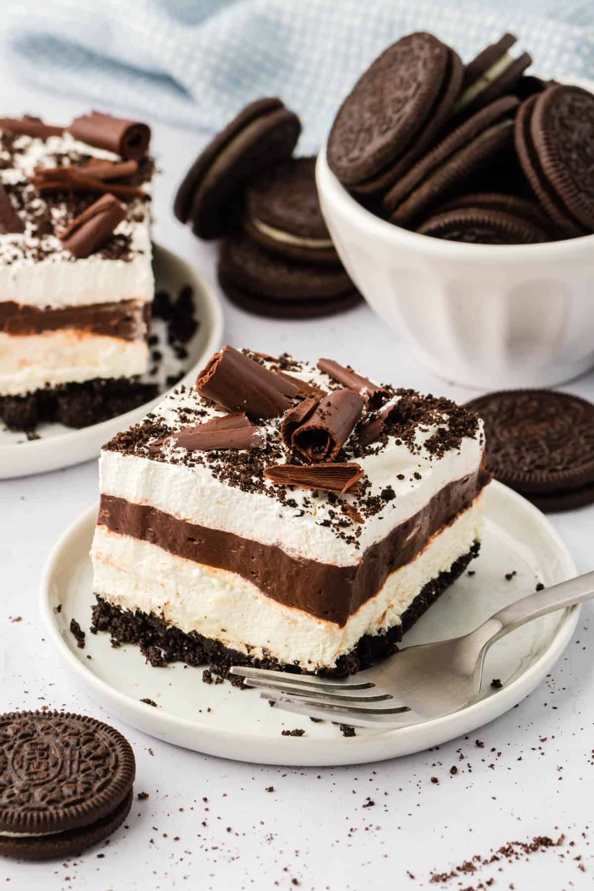 a slice of chocolate lasagna on a white plate surrounded by whole oreos, oreo crumbs, and another slice of lasagna in the background on a plate