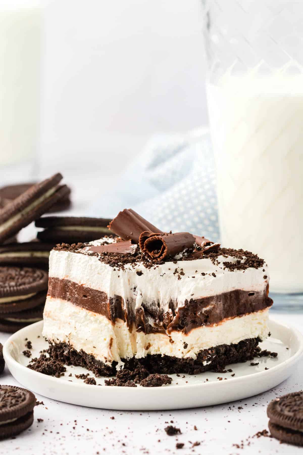 a slice of chocolate lasagna on a white plate with the mark of a fork where one bite has been taken out, surrounded by oreo cookies, cookie crumbs and a glass of milk in the background