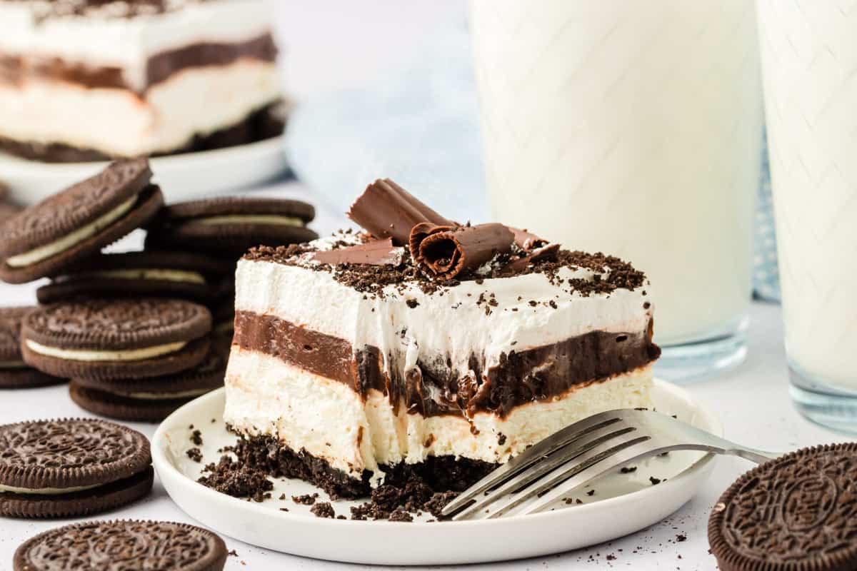 a slice of chocolate lasagna on a white plate with the mark of a fork where one bite has been taken out and a fork on the plate, surrounded by oreo cookies, cookie crumbs and a glass of milk in the background